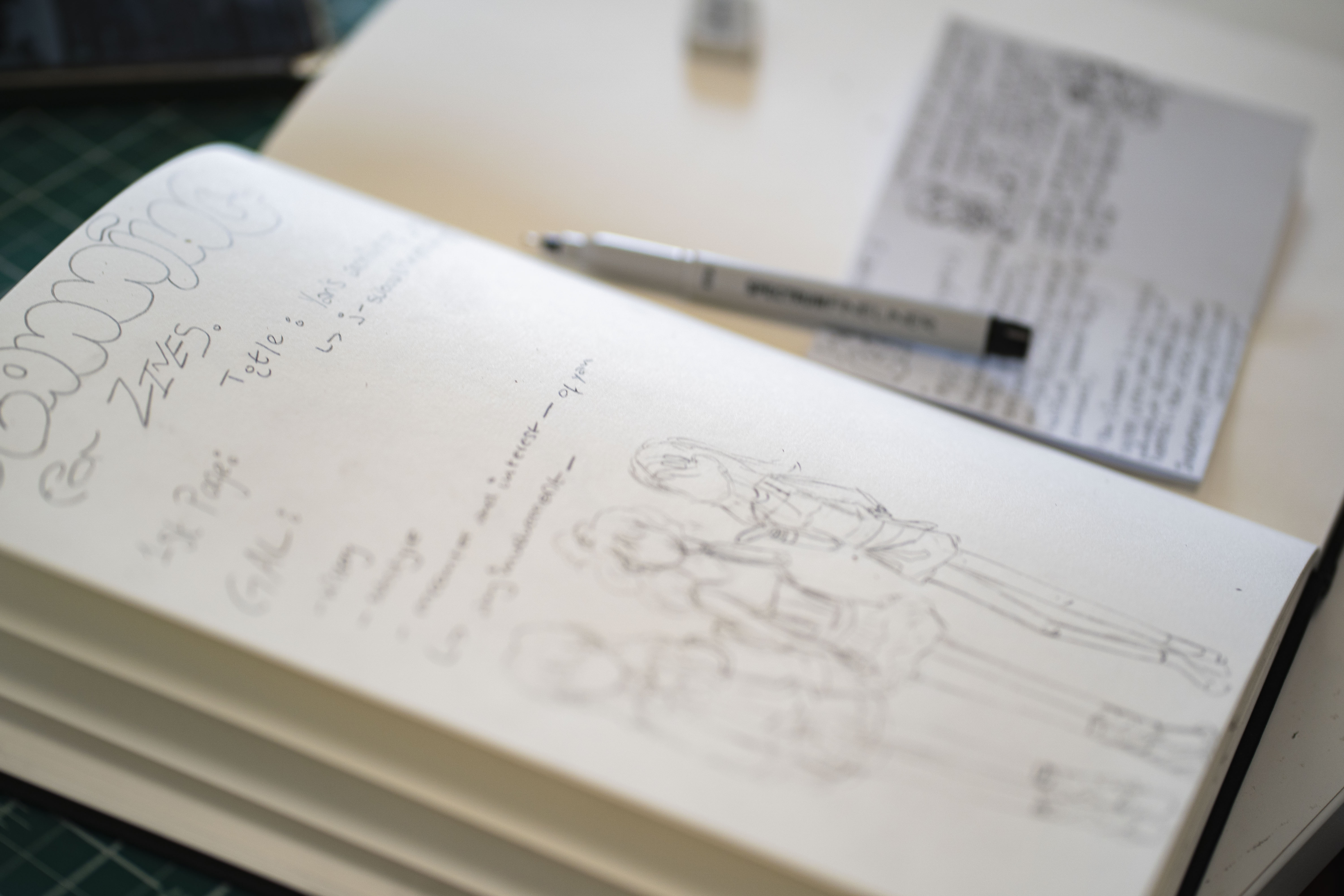 A close-up view of an open sketchbook featuring pencil drawings of three figures on one page. Notes and annotations are visible alongside the sketches. A pen lies across the sketchbook, with another piece of paper partially shown in the background.