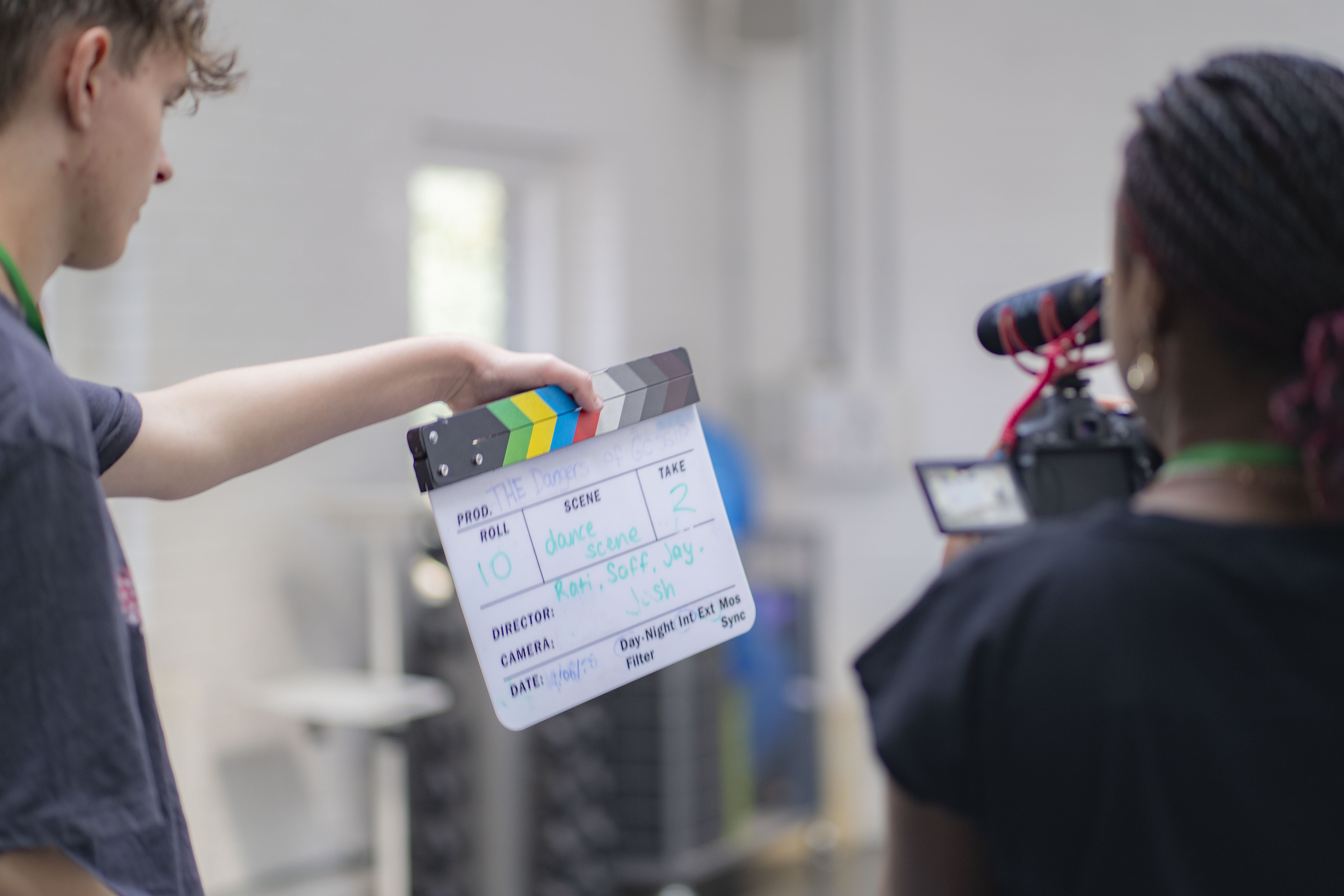 Film crew member holds clapperboard whilst camera operator films in bright indoor location with blurred background.
