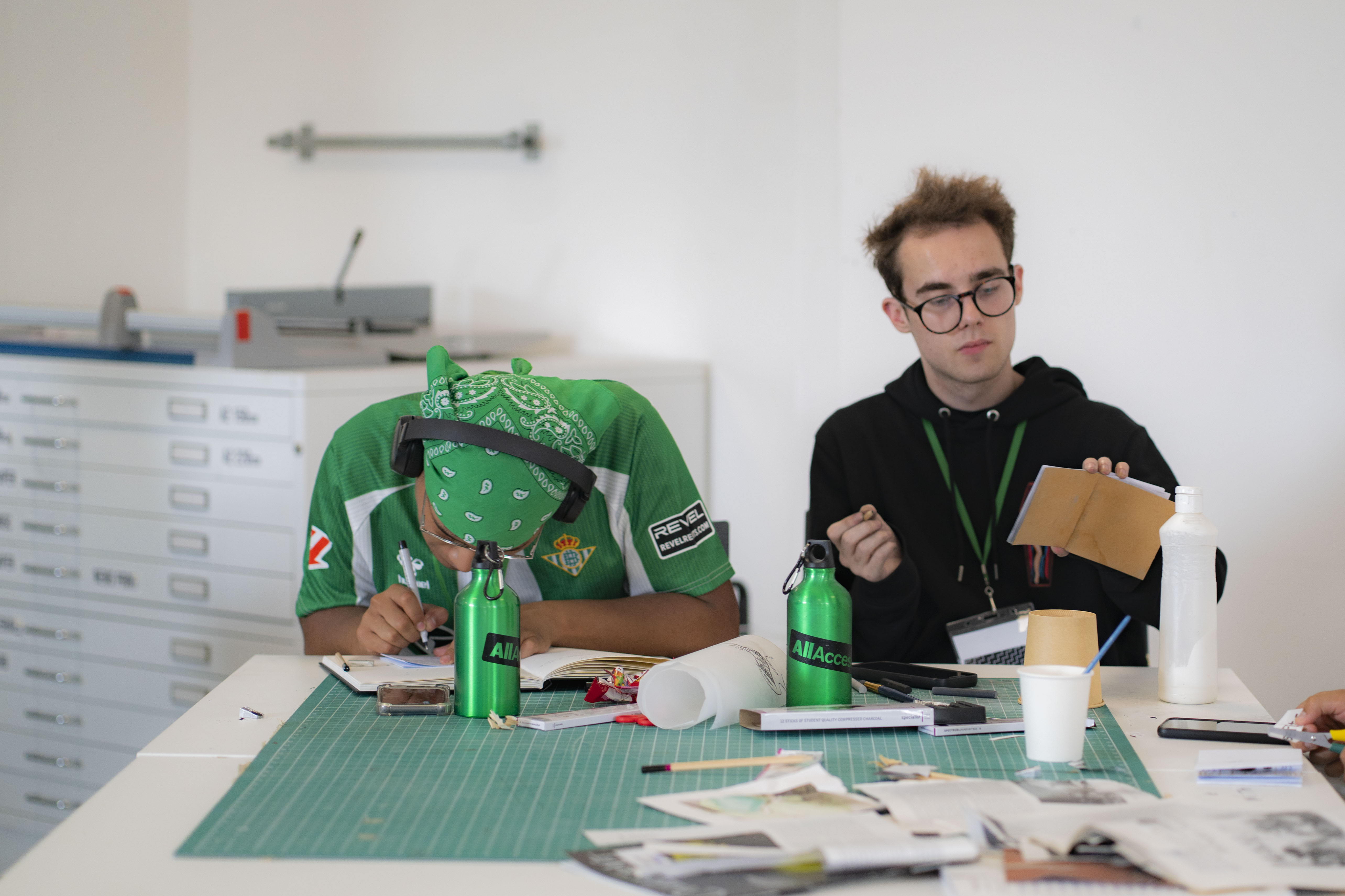 Two people at desk with craft materials. Left person wears bright green hooded jumper, right person in black hoodie with glasses holds brown folder.