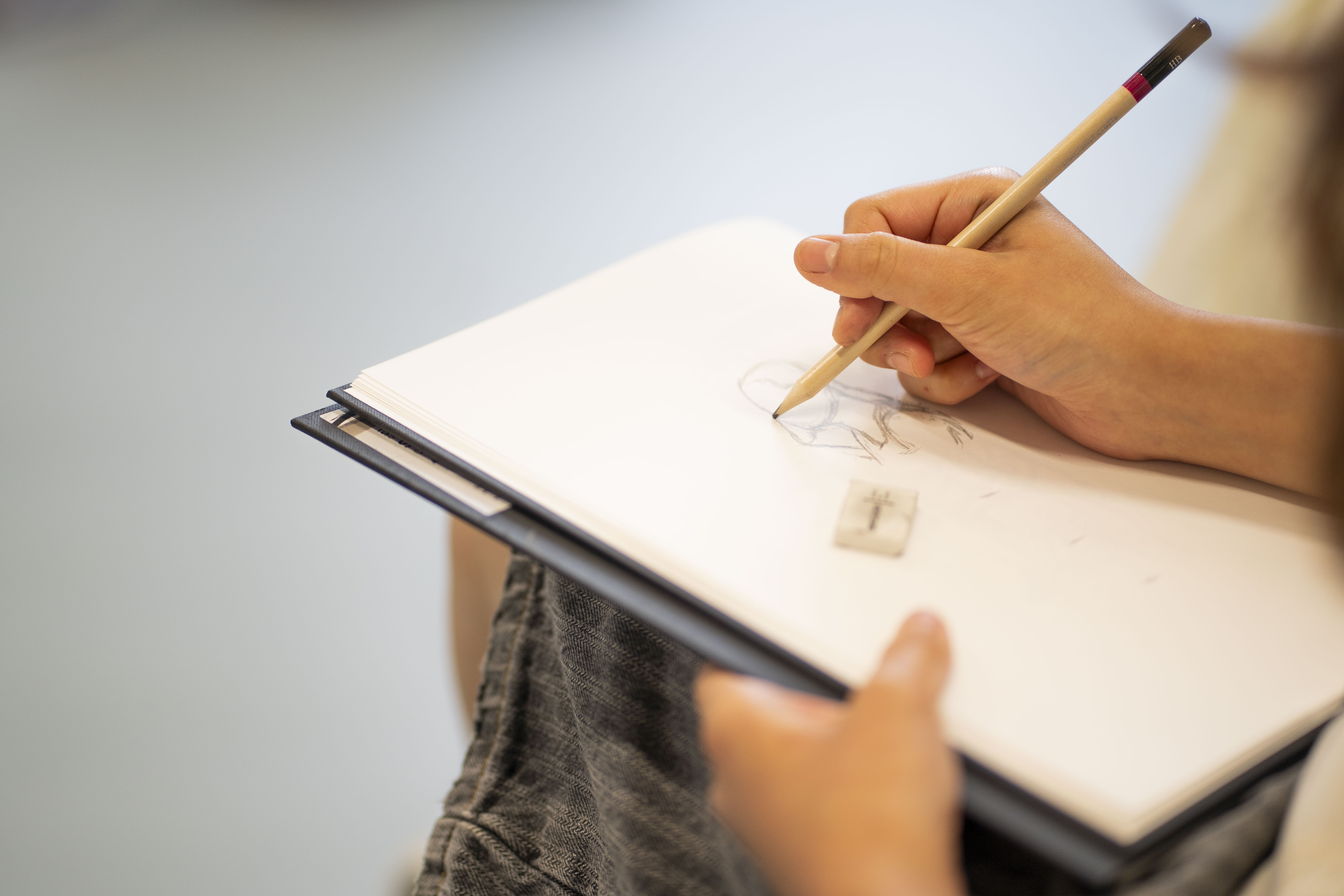 Hand holding pencil drawing on white paper attached to black clipboard, person wearing grey jumper seated.