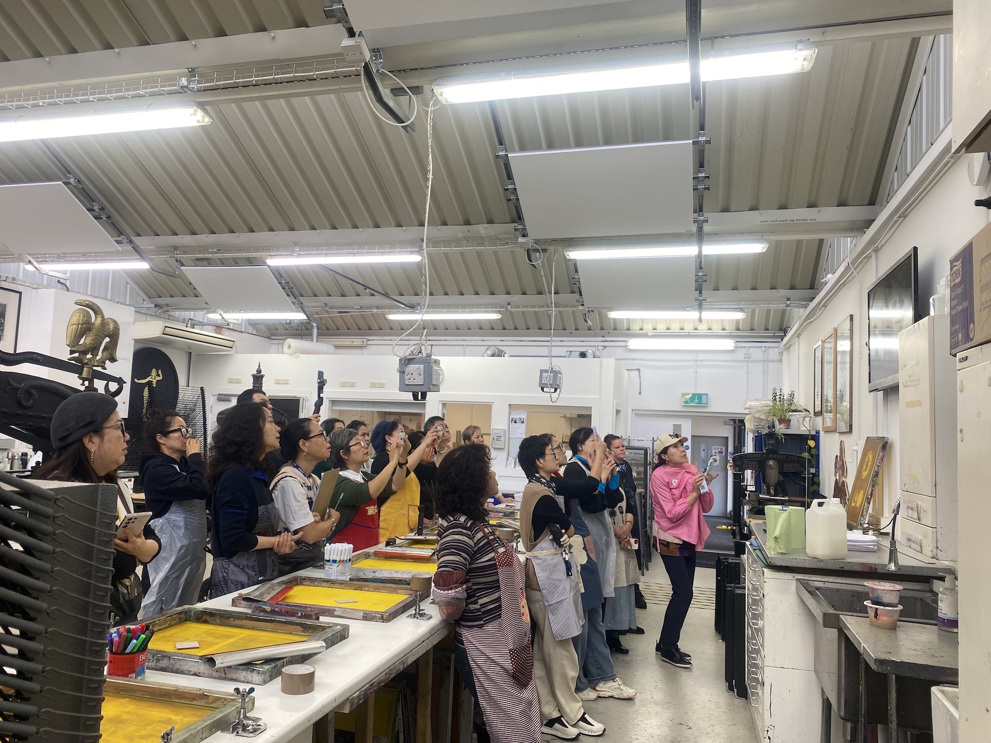 Large group of people standing in industrial workshop with fluorescent lighting, metal ceiling, and yellow work surfaces along the walls.