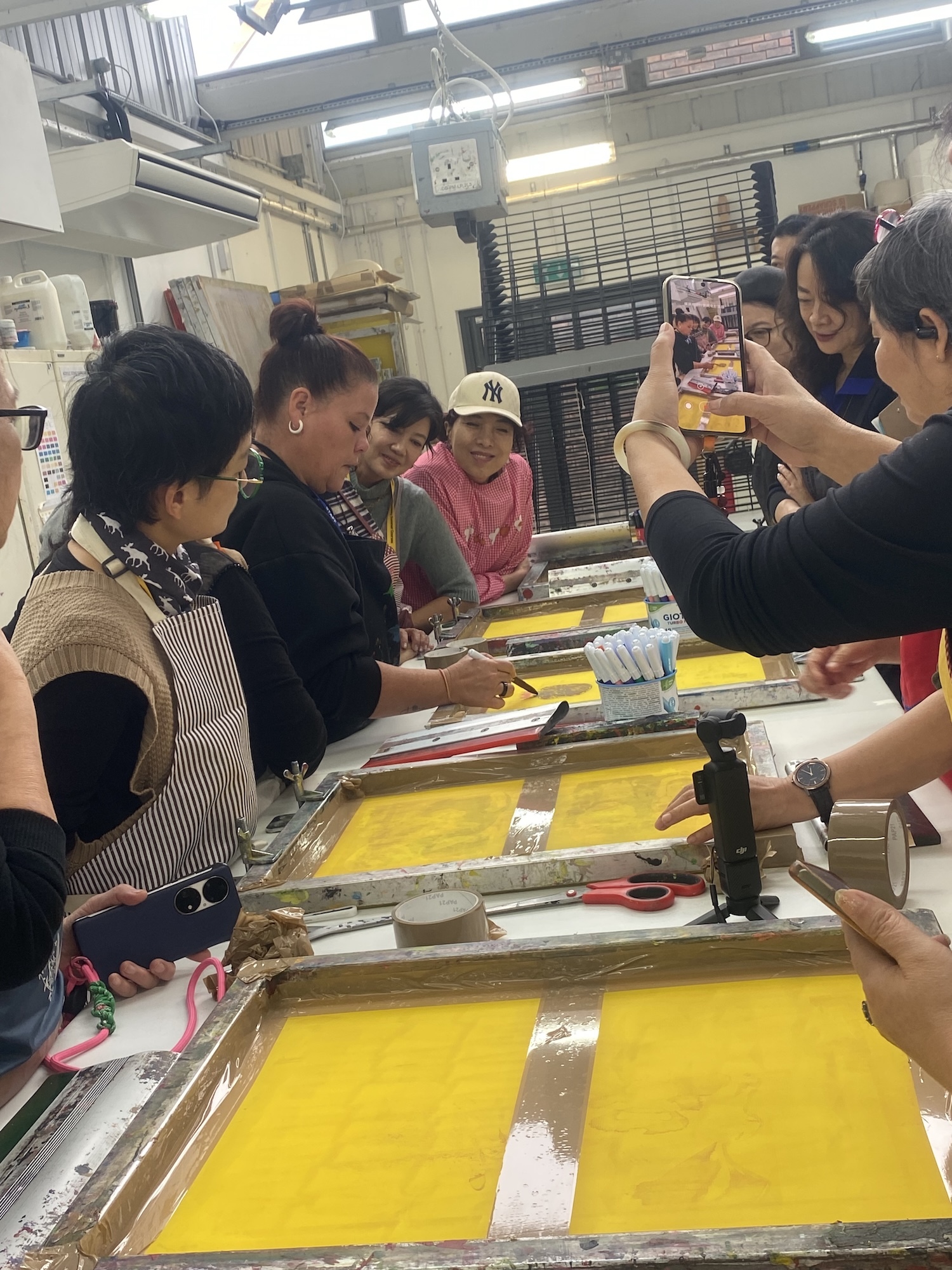 Group of people in printmaking workshop using yellow ink on screens, with one person filming the process on mobile phone.