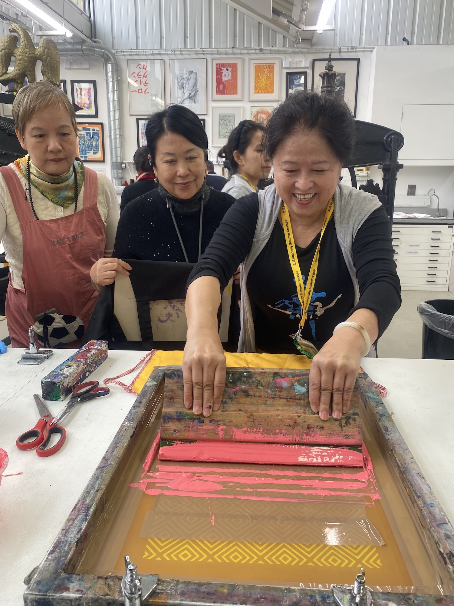 Three women in art studio working on printmaking with red and yellow ink on screen printing frame, framed artwork on white walls.