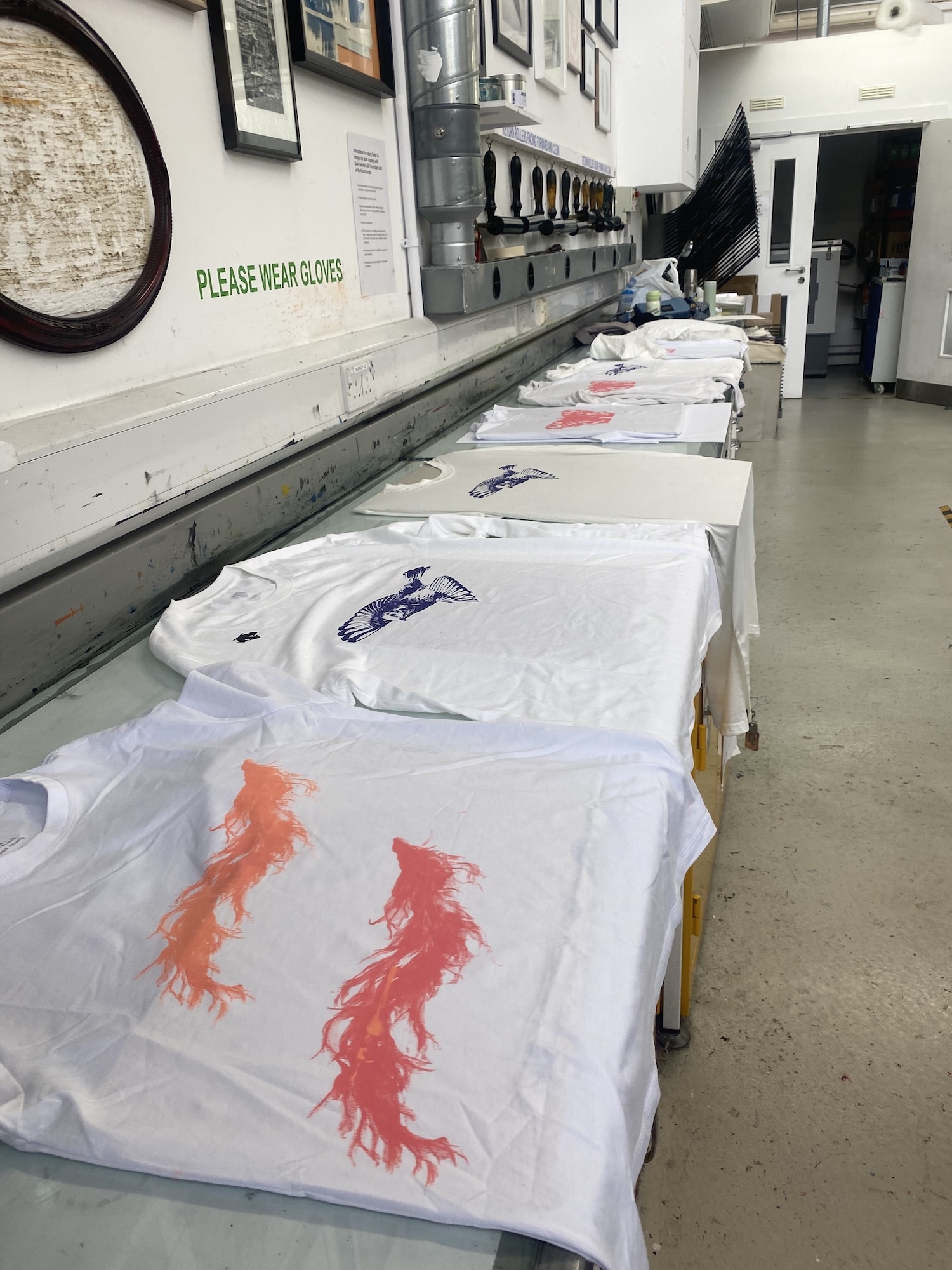 White t-shirts with red and purple designs laid out on tables in screen printing workshop with equipment and framed artwork on walls.