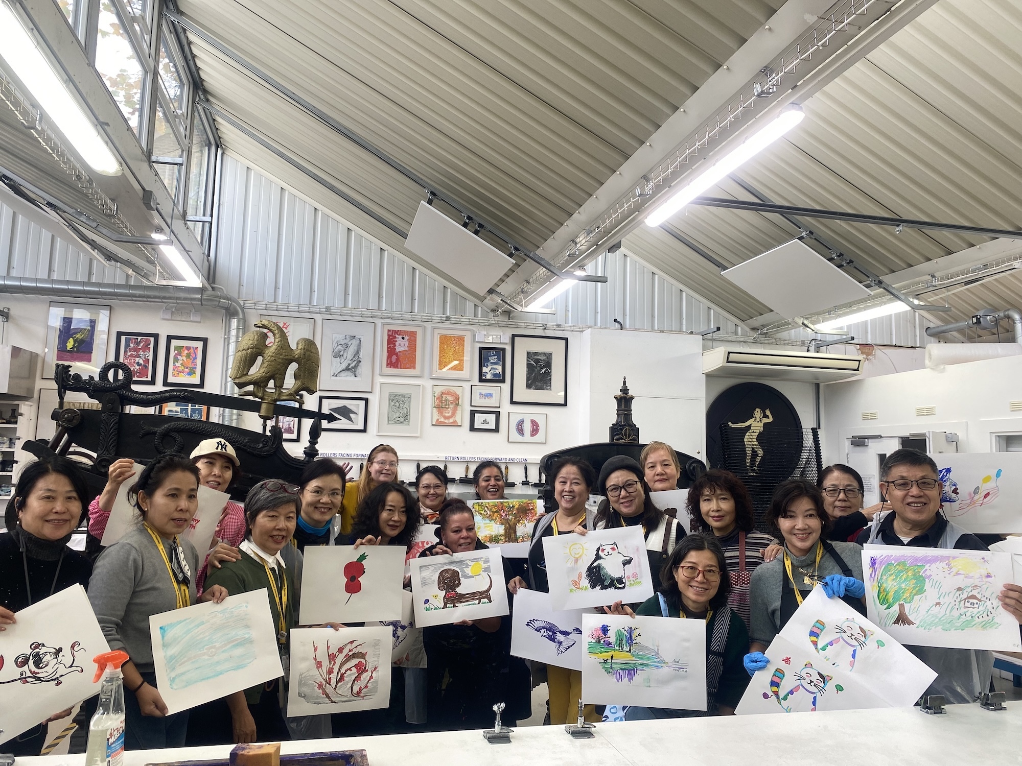 Large group of children and adults in art studio holding up their drawings and paintings, surrounded by framed artwork on walls.