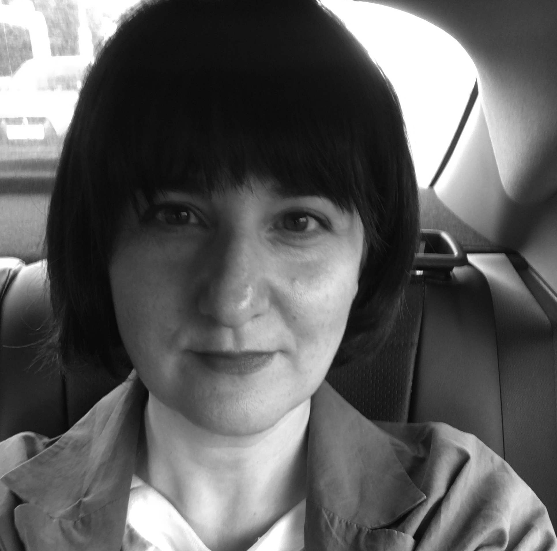 Woman with dark bob haircut and fringe sitting in car, wearing collared shirt. Black and white image with car interior visible.