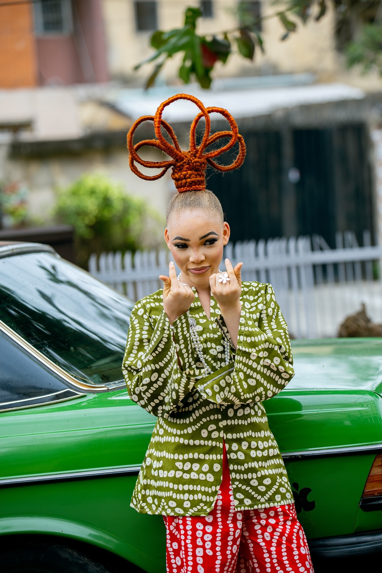 Woman with orange rope headpiece making peace signs, wearing green patterned top and red bottoms, standing by green car.