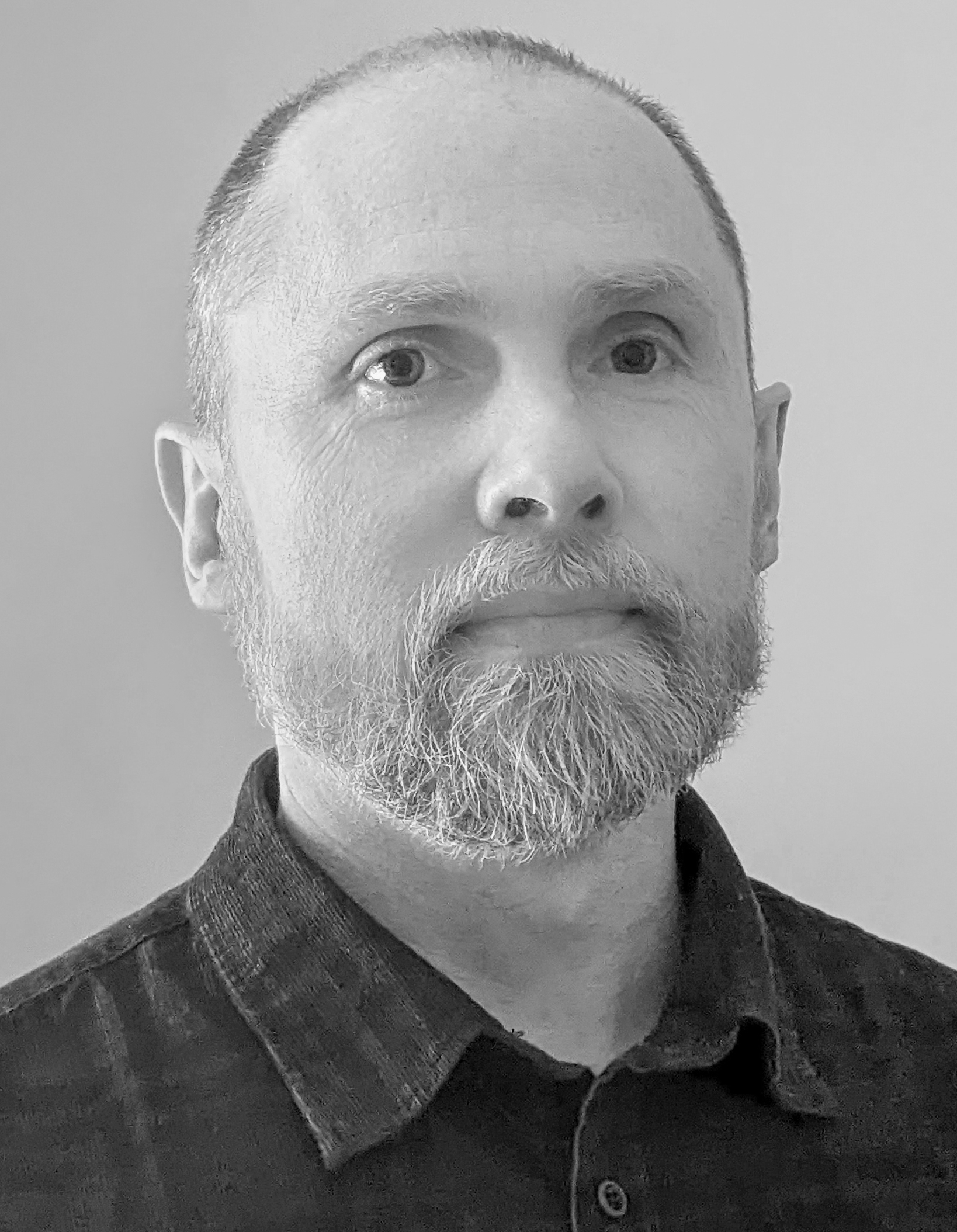 Middle-aged man with grey beard and receding hairline wearing dark collared shirt, black and white portrait against plain background.