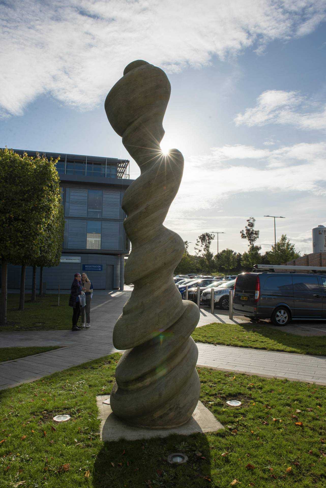 Abstract sculpture with the sun behind it and a university building in the background.