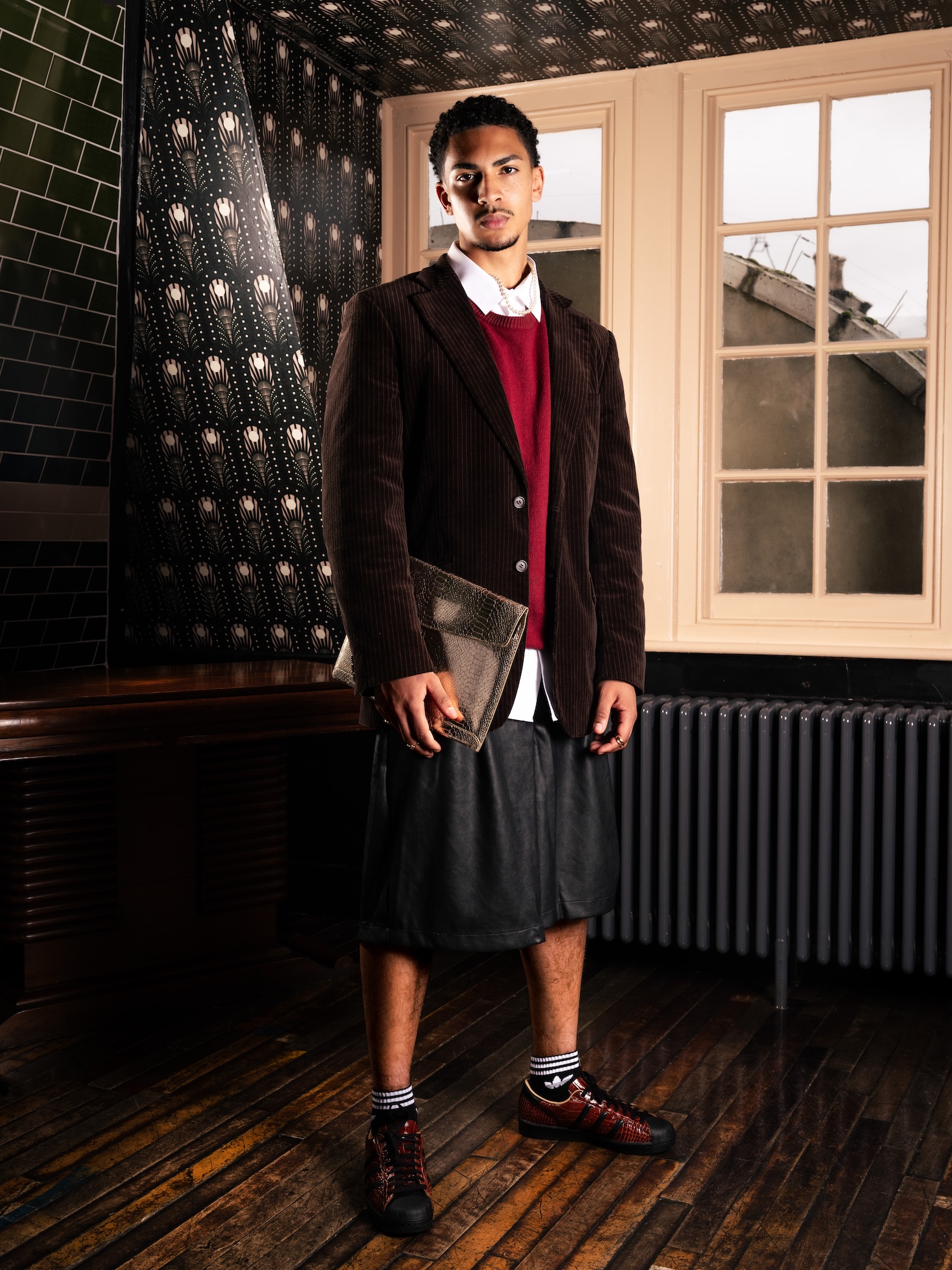 A man stands in a room with patterned wallpaper and large windows. He wears a brown cord jacket over a red sweater, a white shirt, and a black skirt. He holds a laptop case and wears red and black shoes with striped socks.