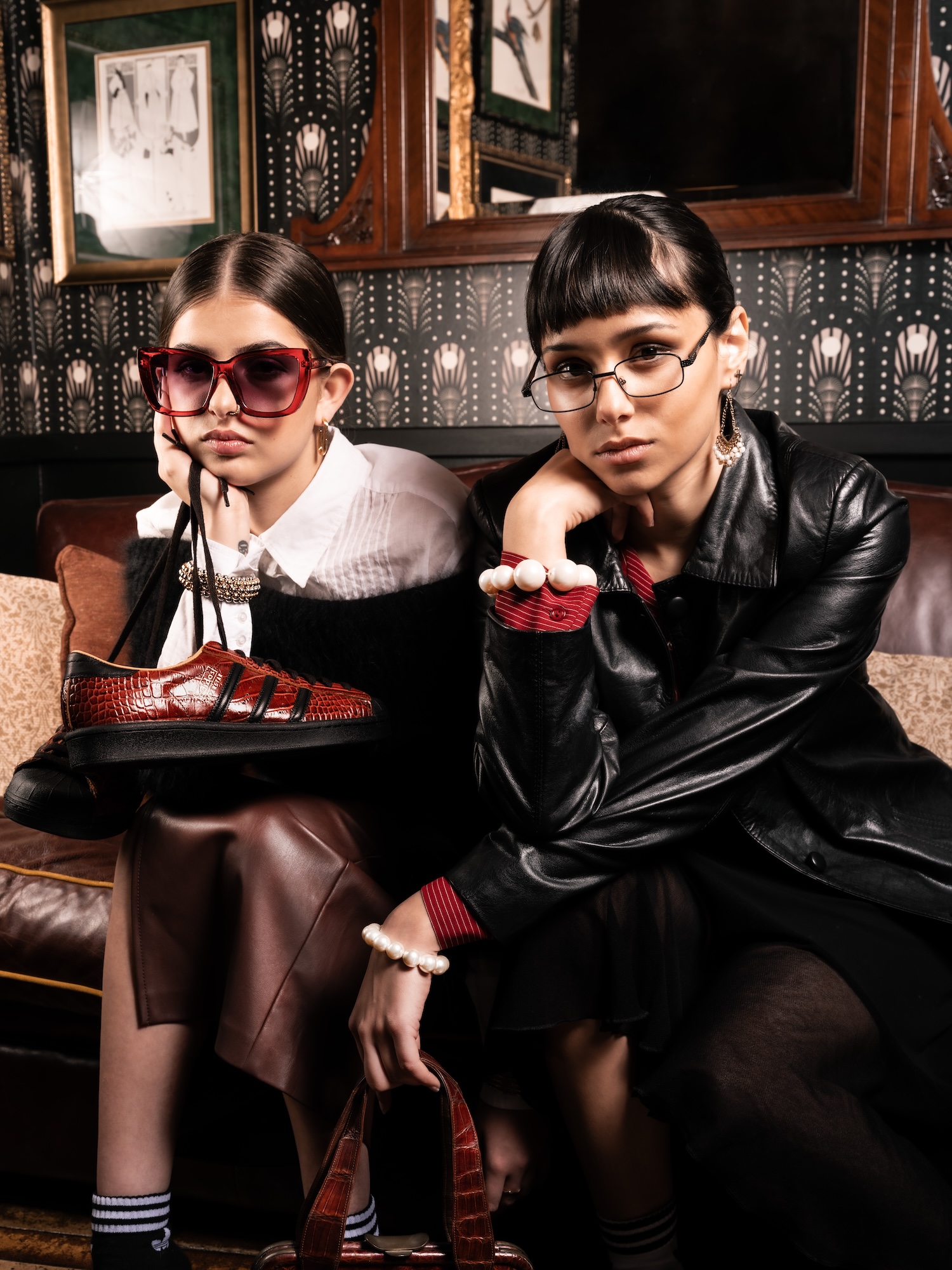 Two young women pose on a sofa in a stylish interior. One woman, wearing oversized pink sunglasses, holds a pair of black and red shoes. The other woman, dressed in a black leather jacket, rests her chin on her hand. The background features patterned wallpaper and framed pictures.