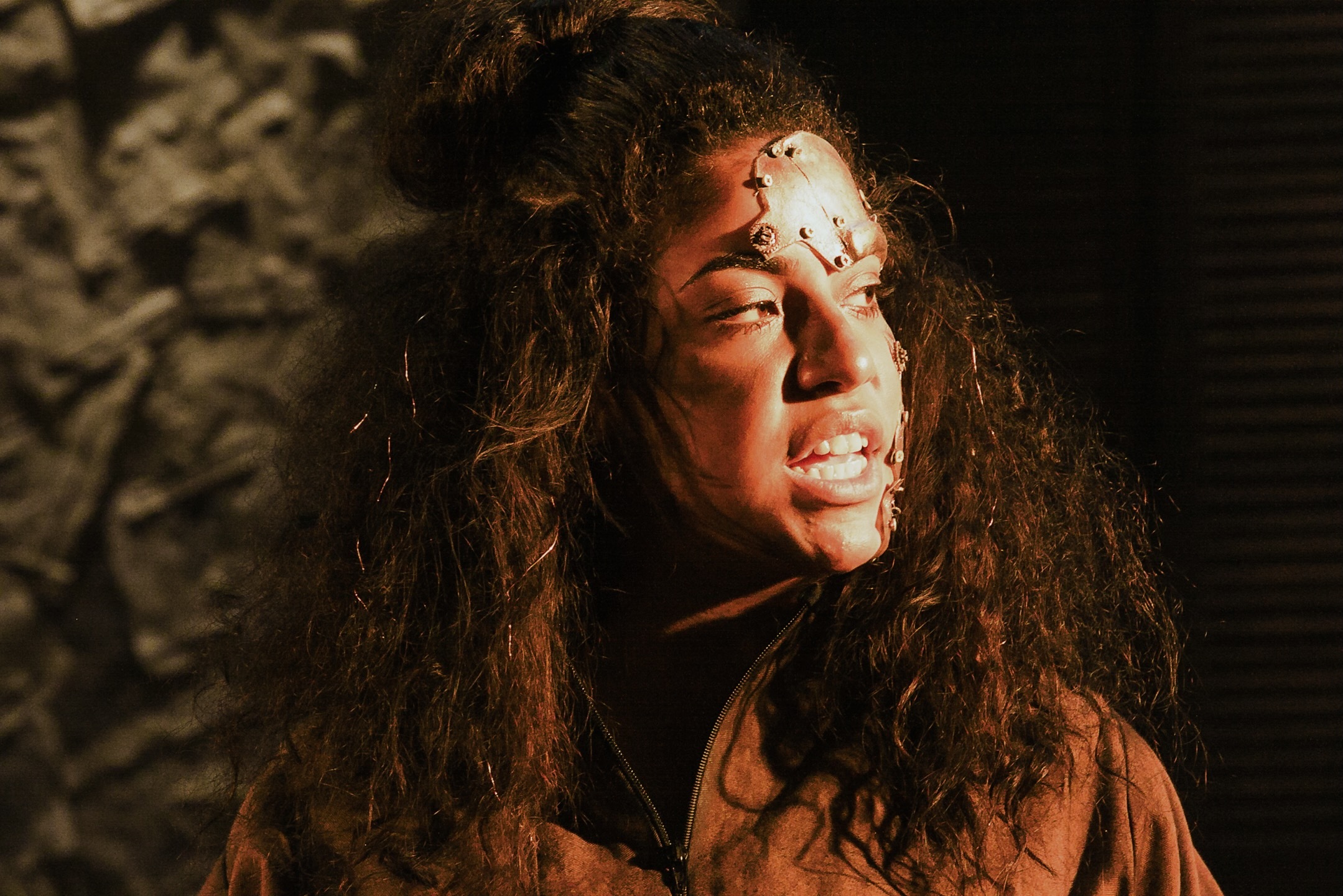 Young woman with long curly hair and bandage on forehead, eyes closed, warm light illuminating face against dark background.
