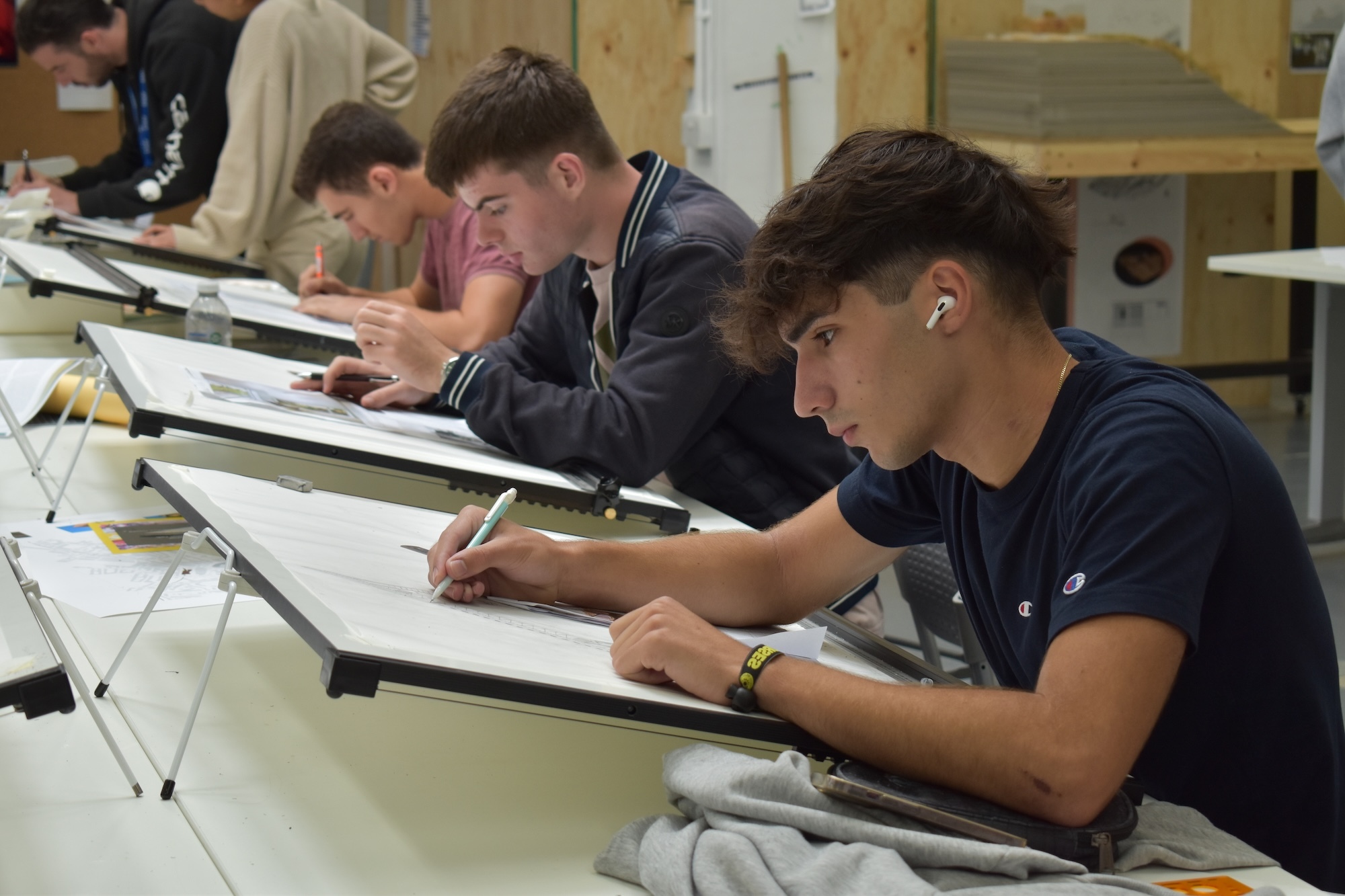 Students sitting at angled drawing boards in classroom, writing or drawing on paper with pencils, wearing dark clothing.