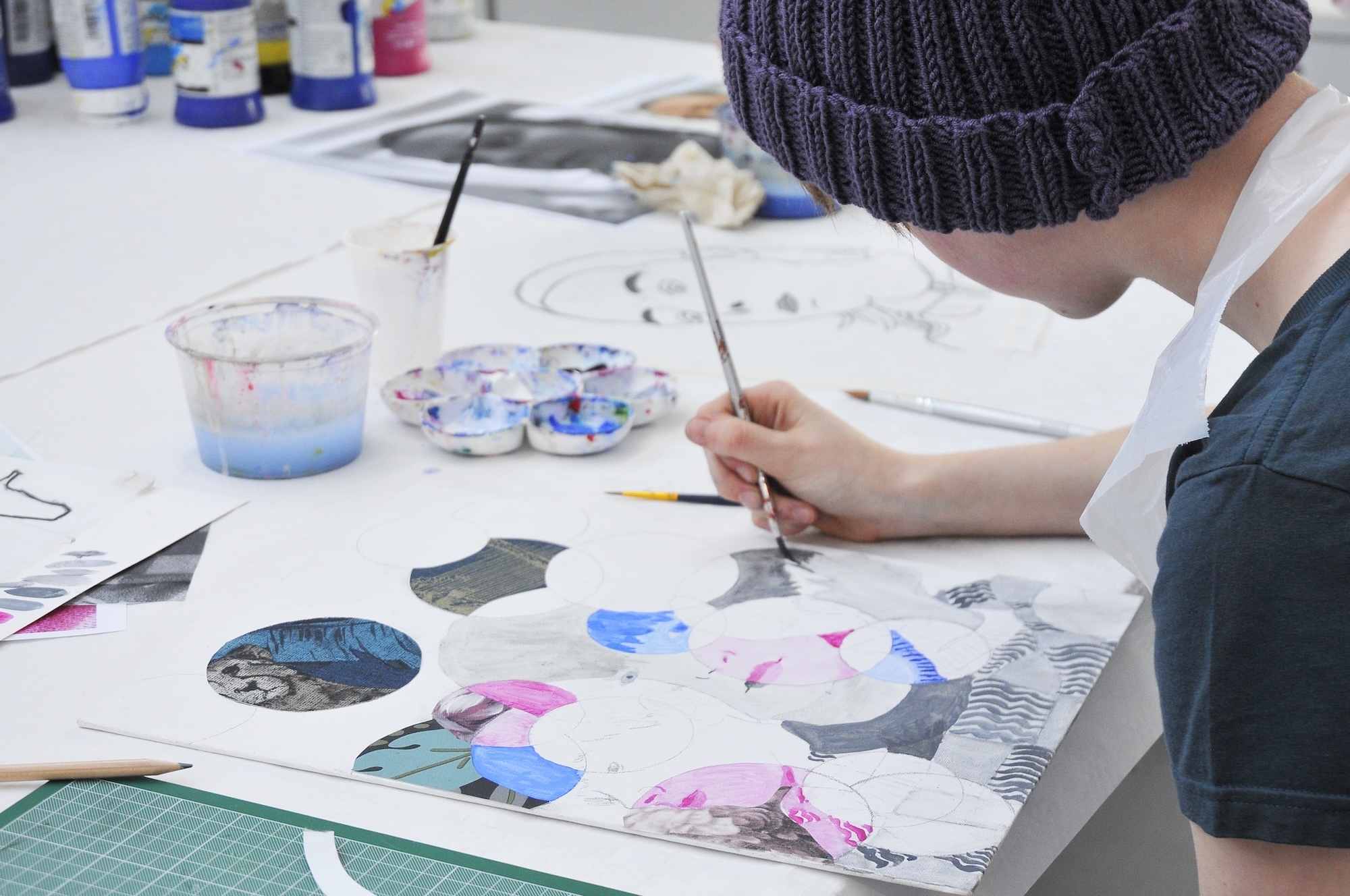 Person in grey knitted hat painting watercolours on white paper at desk with palette, brushes, and water jar visible.