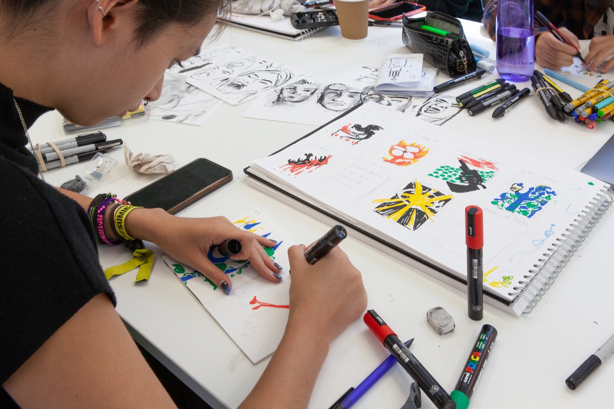 Person drawing with coloured markers at white table covered in art supplies, sketches, and colourful artwork samples.