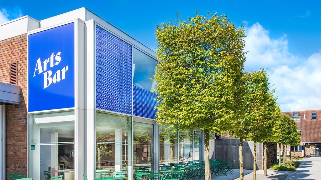A row of trees trimmed to resemble cubes outside a university building with outdoor seating. The words 'Arts Bar' appears on the outside of the building.