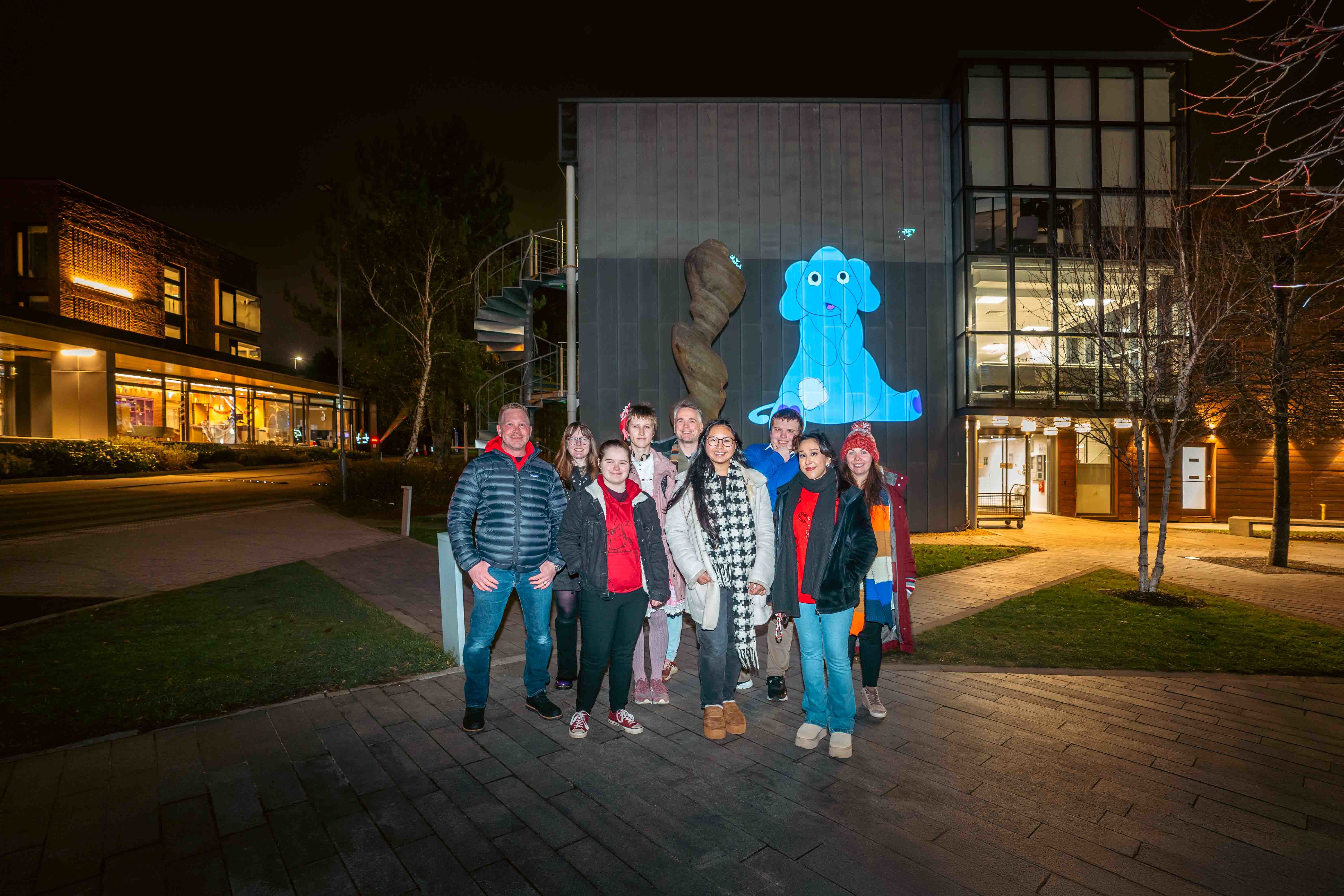 A group of AUB students and community members gathered outside University House on AUB campus at night posing for a photo. A blue animated elephant is projected onto the front of the building behind them.