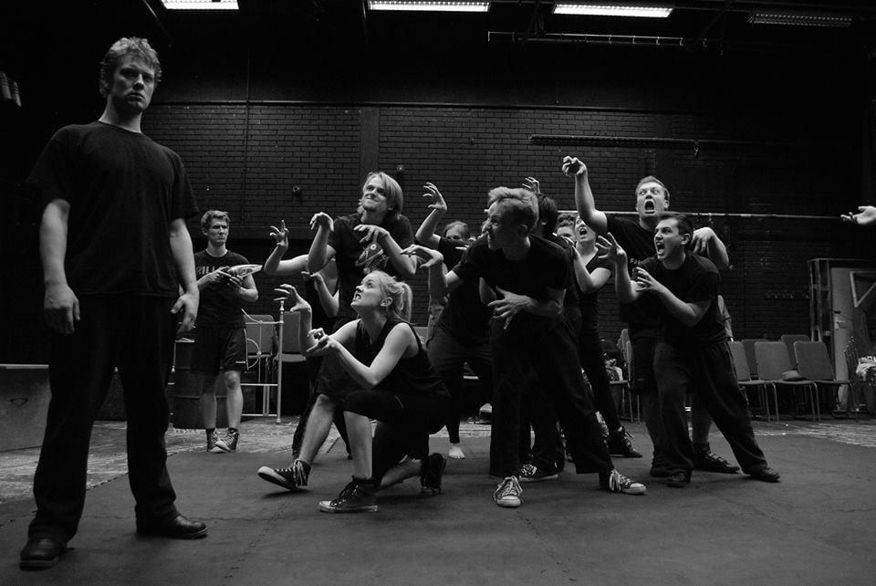A performer stands with a serious expression on the left-hand side with a group of other performers reaching towards him.