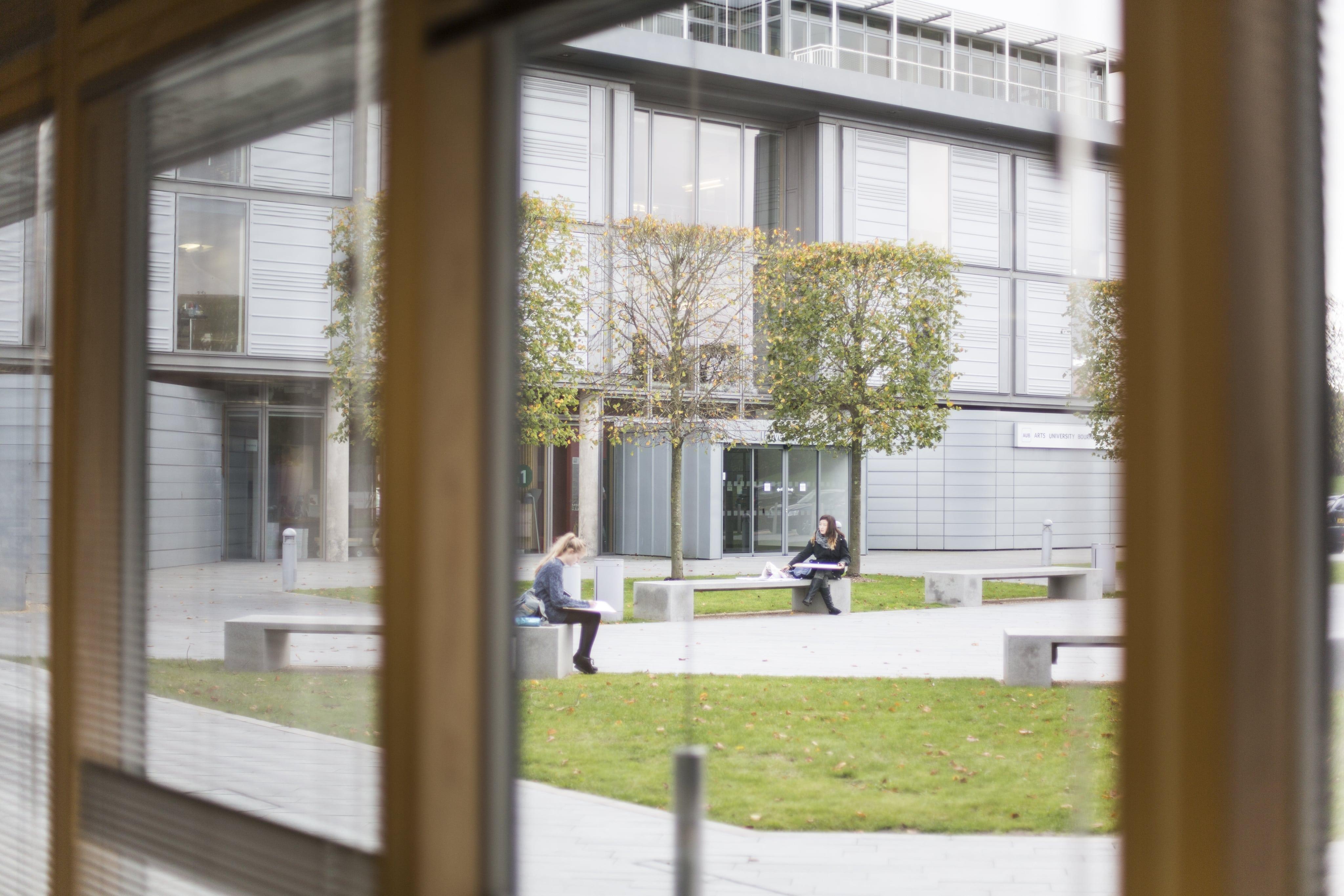 A photograph taken through a window showing a university building and a few students sitting on benches.
