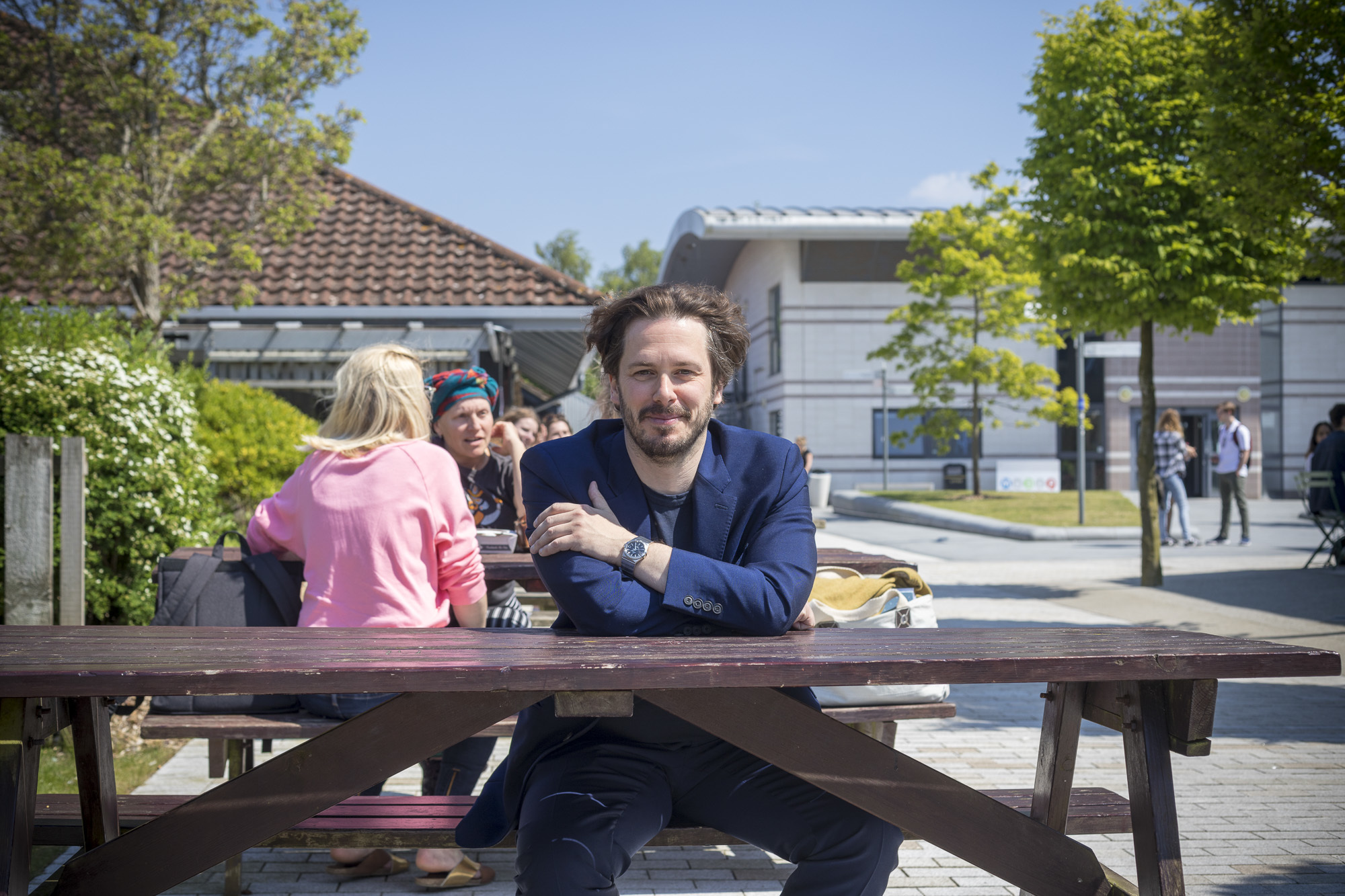 Edgar Wright visits AUB. He is sat in the centre of a picnic table on AUB's campus. Two other people sit behind him at another bench.