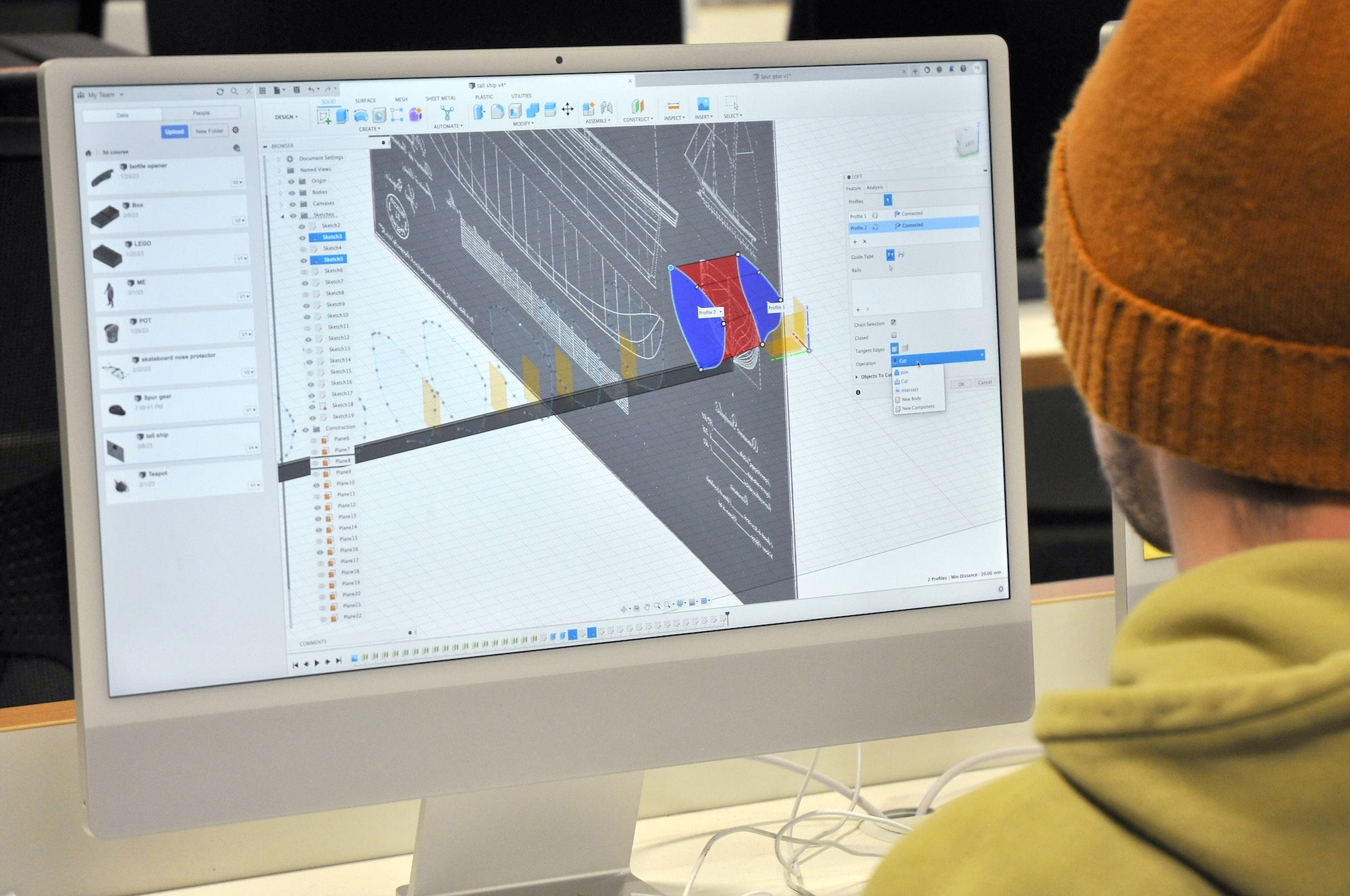Person in orange beanie and yellow hoodie viewing computer monitor displaying 3D CAD software with blue and red geometric model.