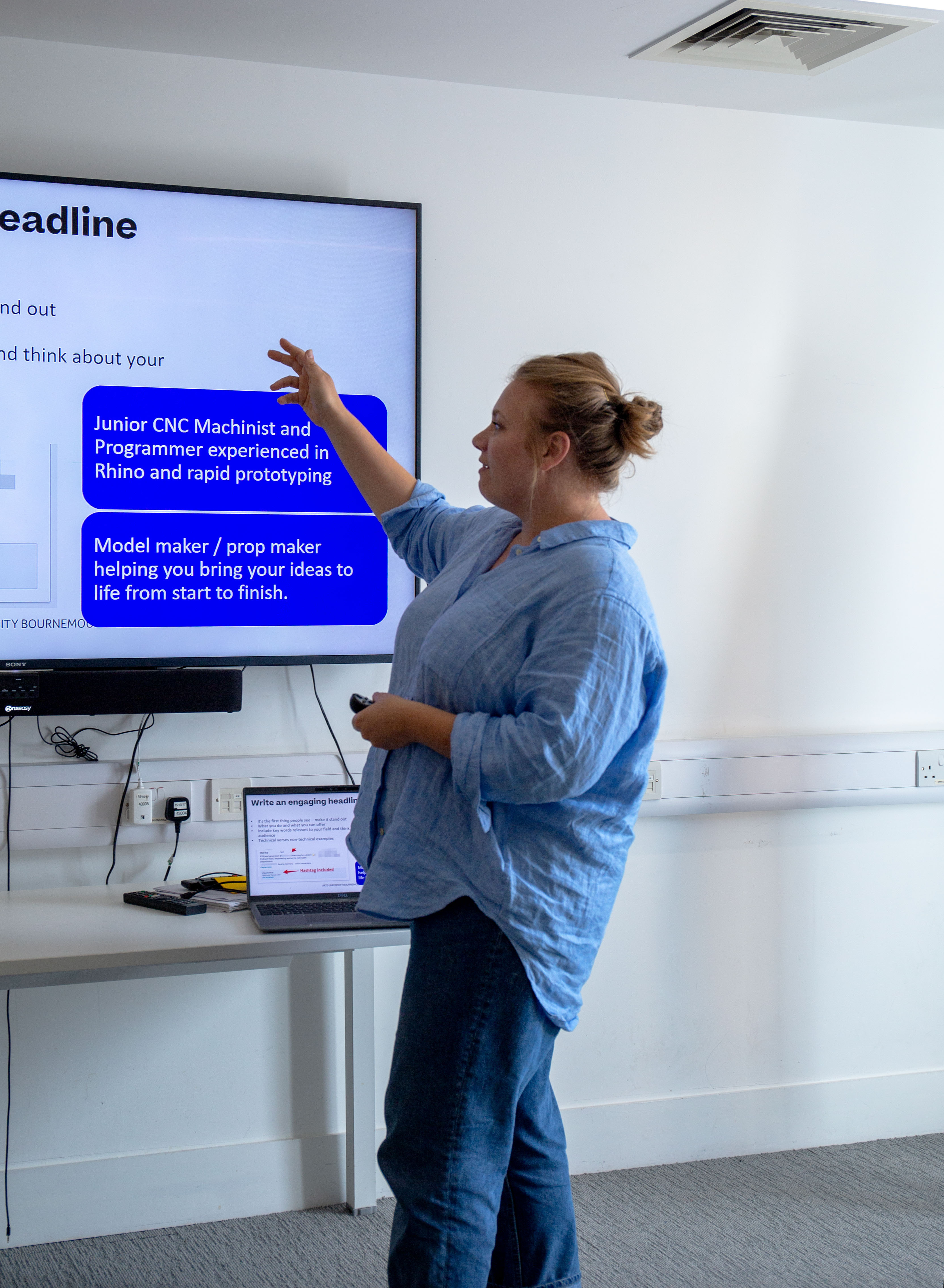Man in blue shirt pointing at wall-mounted screen displaying job advert text, standing beside laptop on white desk in office room.
