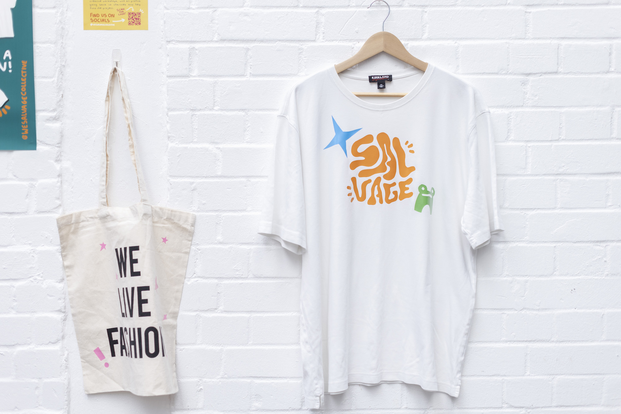 A tote bag and a white T-shirt with the Salvage Collective logo on a hanger hang on display on a white brick wall.