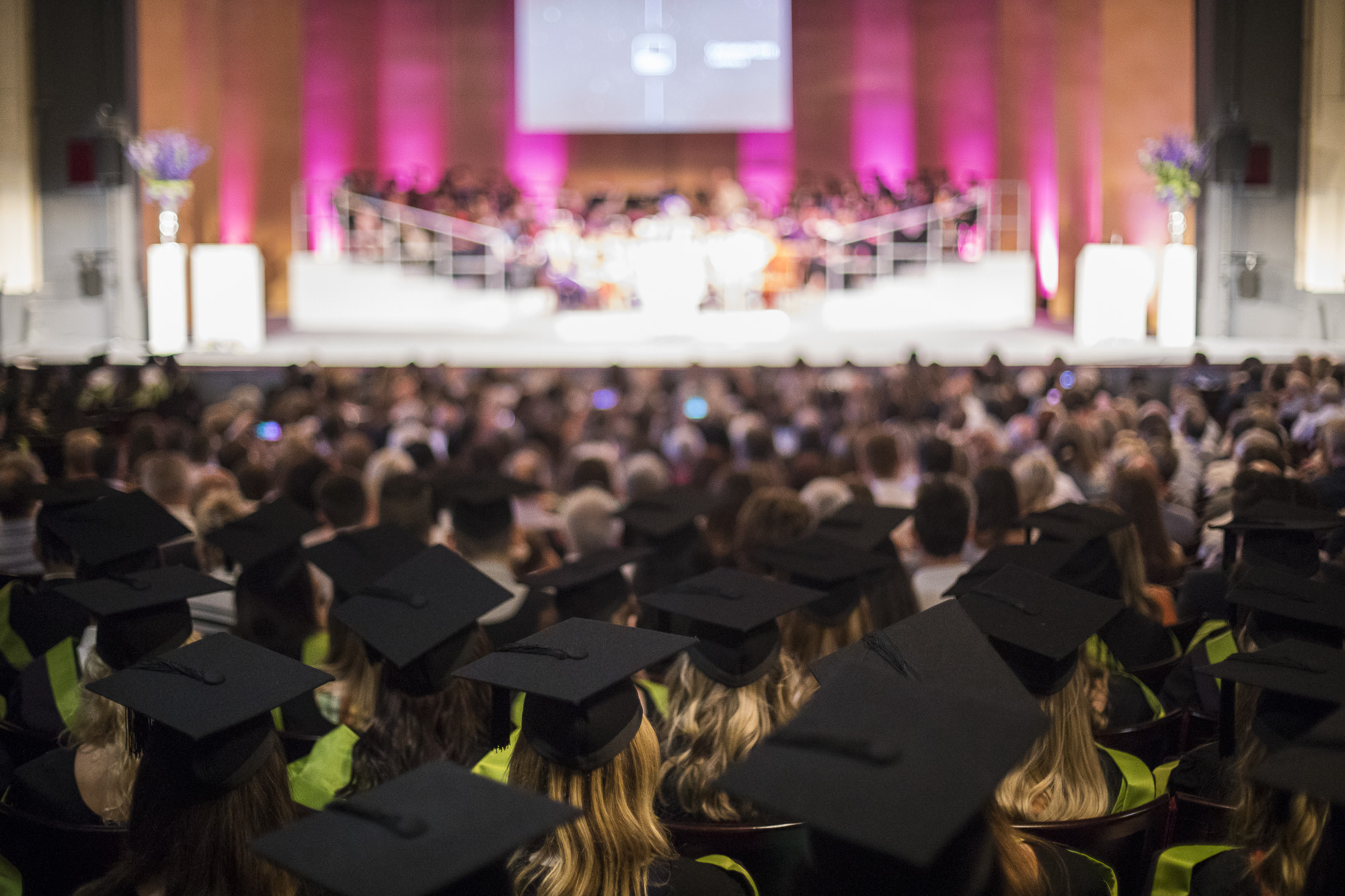 A theatre full of people in graduation dress, viewed form the back with a stage blurred in the background.