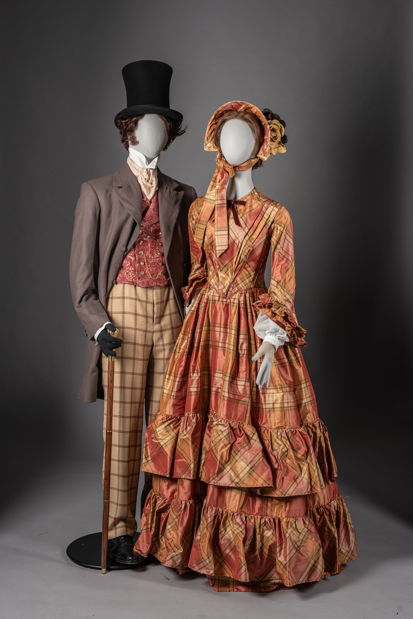 Two mannequins dressed in 19th century Wuthering Heights-inspired costumes. One with a men's frock coat, waistcoat, trousers and top hat and holding a cane; the other wearing an orange patterned day dress and bonnet.