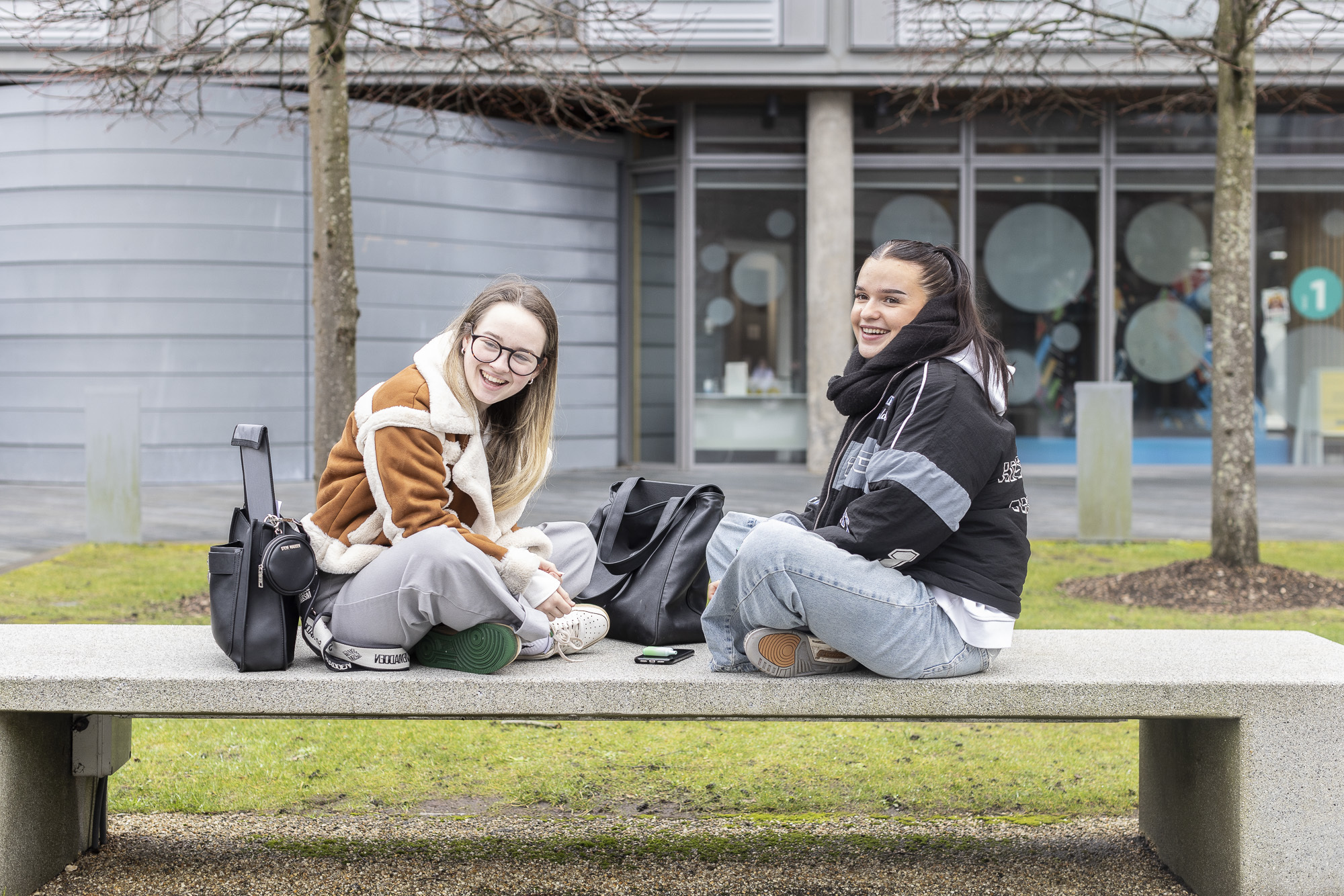 Two AUB students sitting opposite each other smiling on a bench in the courtyard outside South House, AUB Campus.