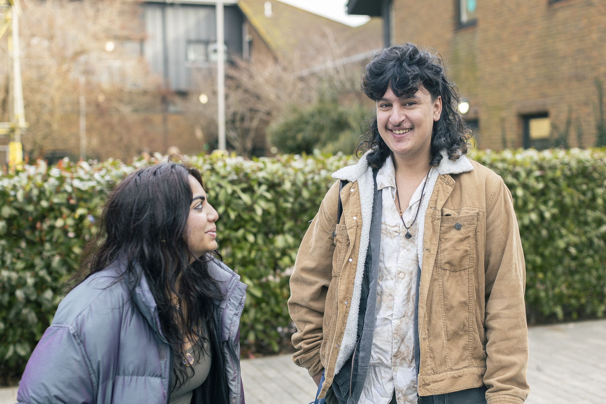 Two Arts University Bournemouth students on campus, one is sitting and looking up at the other, who is looking at the camera and smiling.
