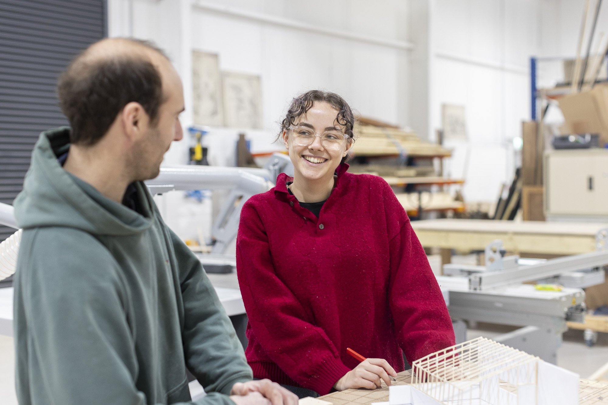 Two people in workshop setting - member of Arts University Bournemouth staff in grey hoodie and an Arts University Bournemouth student in red jumper and glasses, smiling whilst chatting.