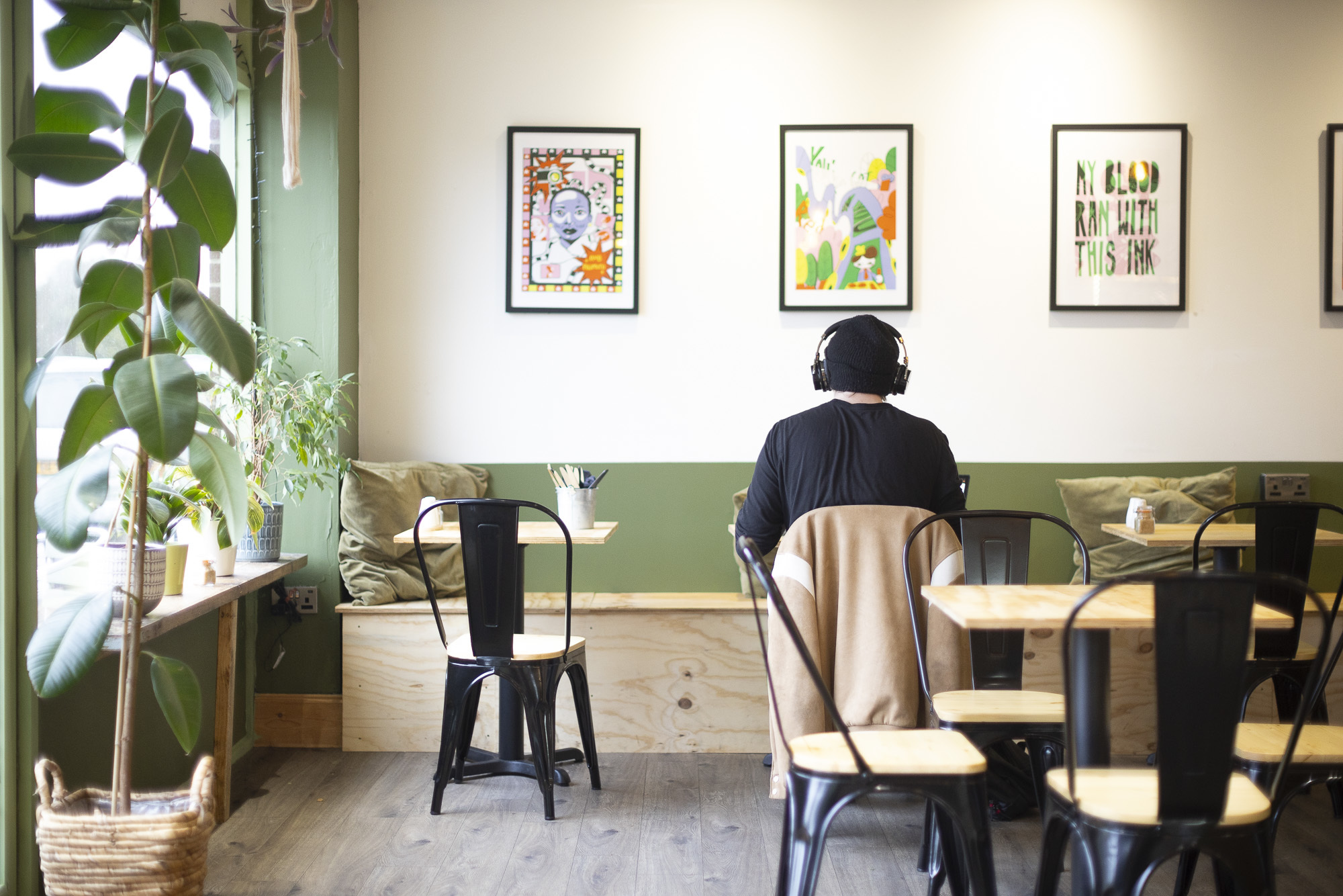 A person sitting with headphones on, with their back to the camera, in a coffee shop, with three framed artworks on the wall above them.