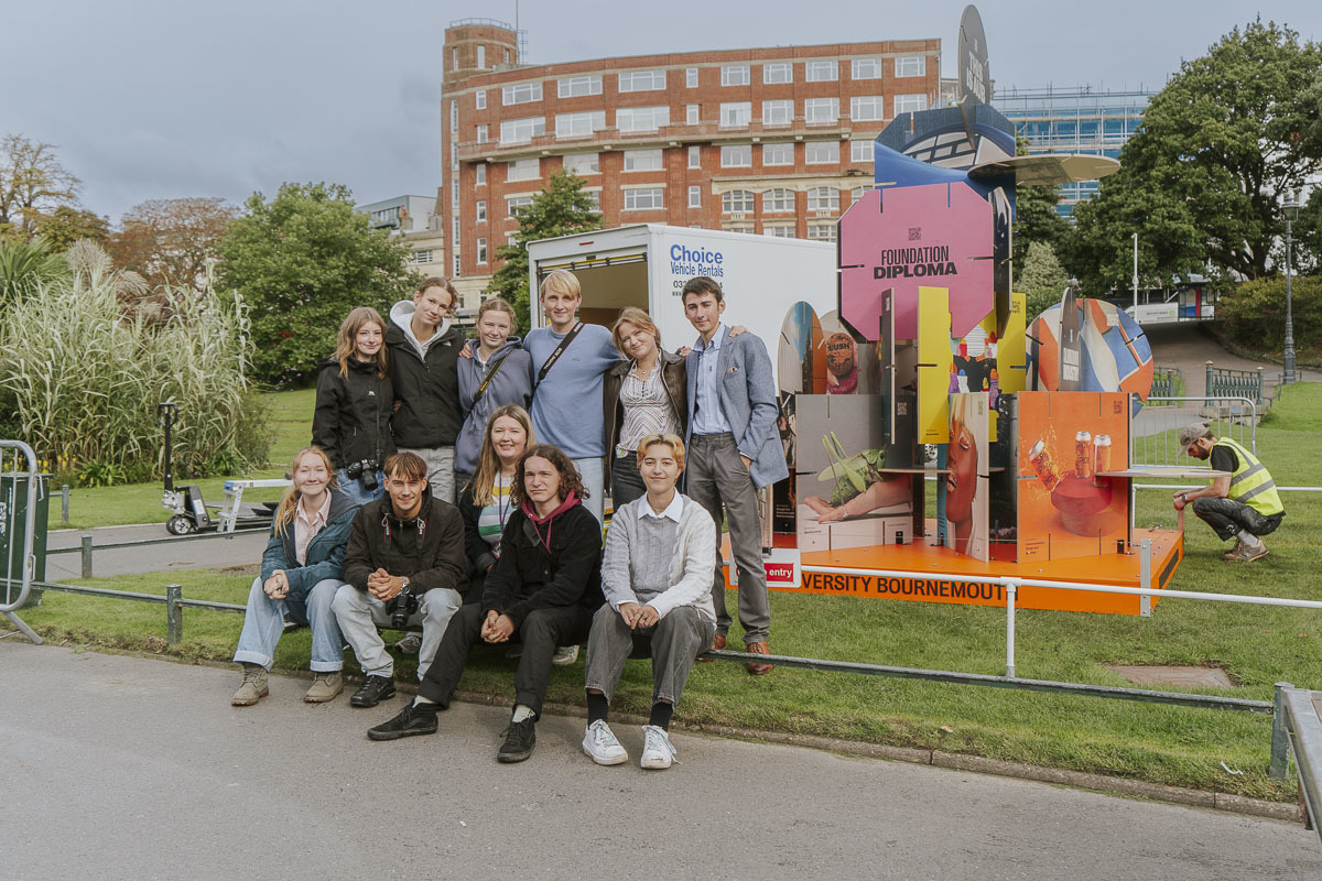 A group of eleven young people gathered and posing for a photo next to a sculpture based on AUB's House of Cards prospectus in Bournemouth Gardens.
