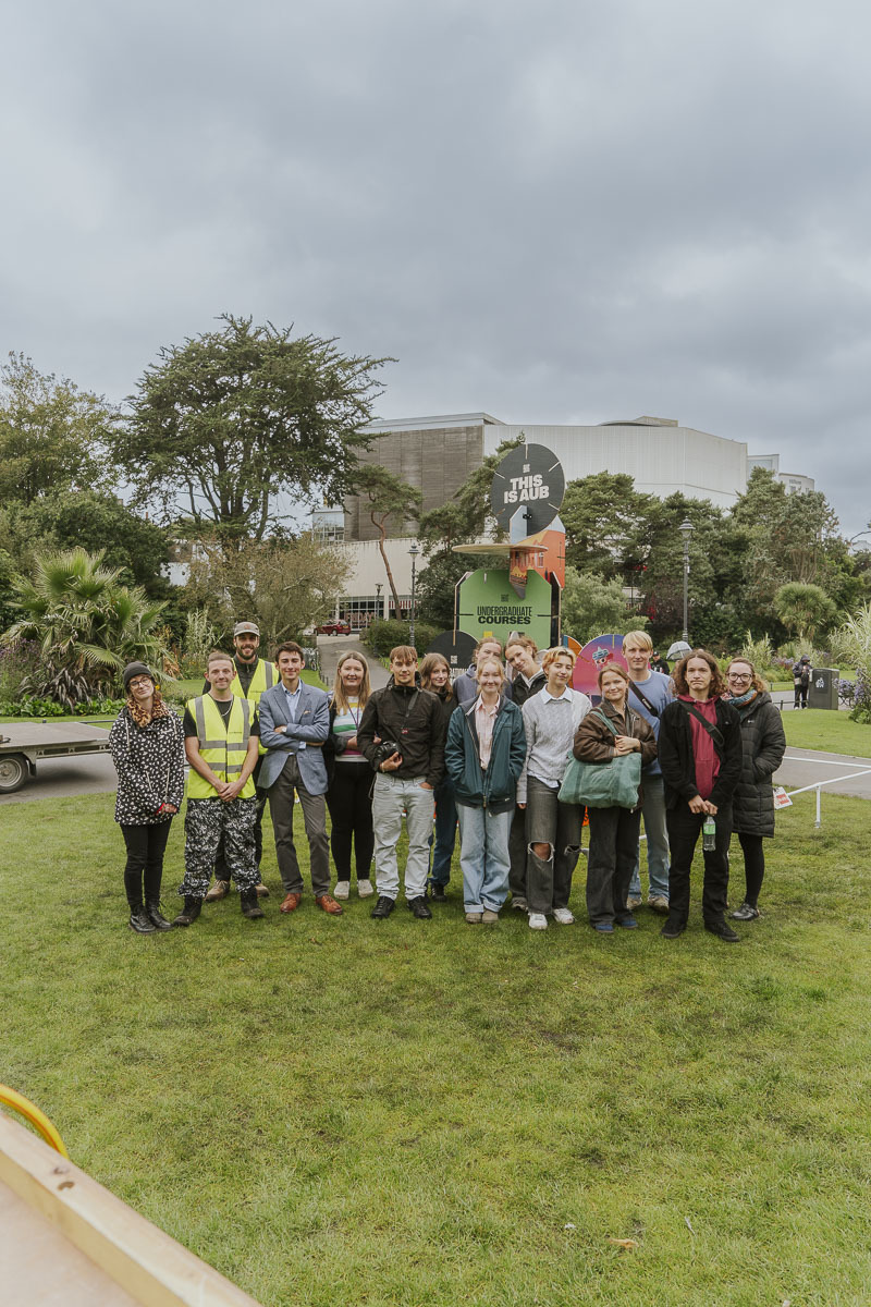 A large group of people gathered and posing for a photo in front of a large sculpture based on AUB's House of Cards prospectus in Bournemouth Gardens.