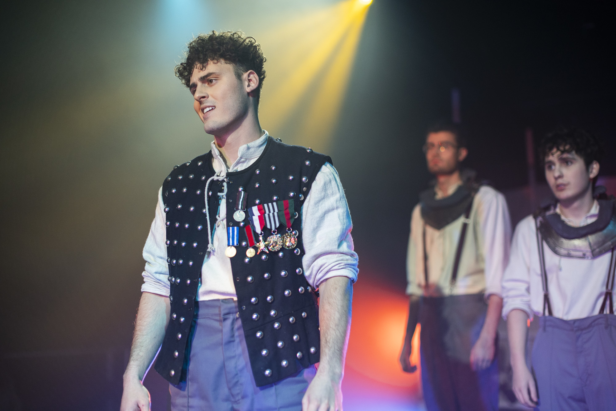A BA (Hons) Acting student wearing an open waistcoat with several medals on the right-hand side. Two fellow students stand behind him.