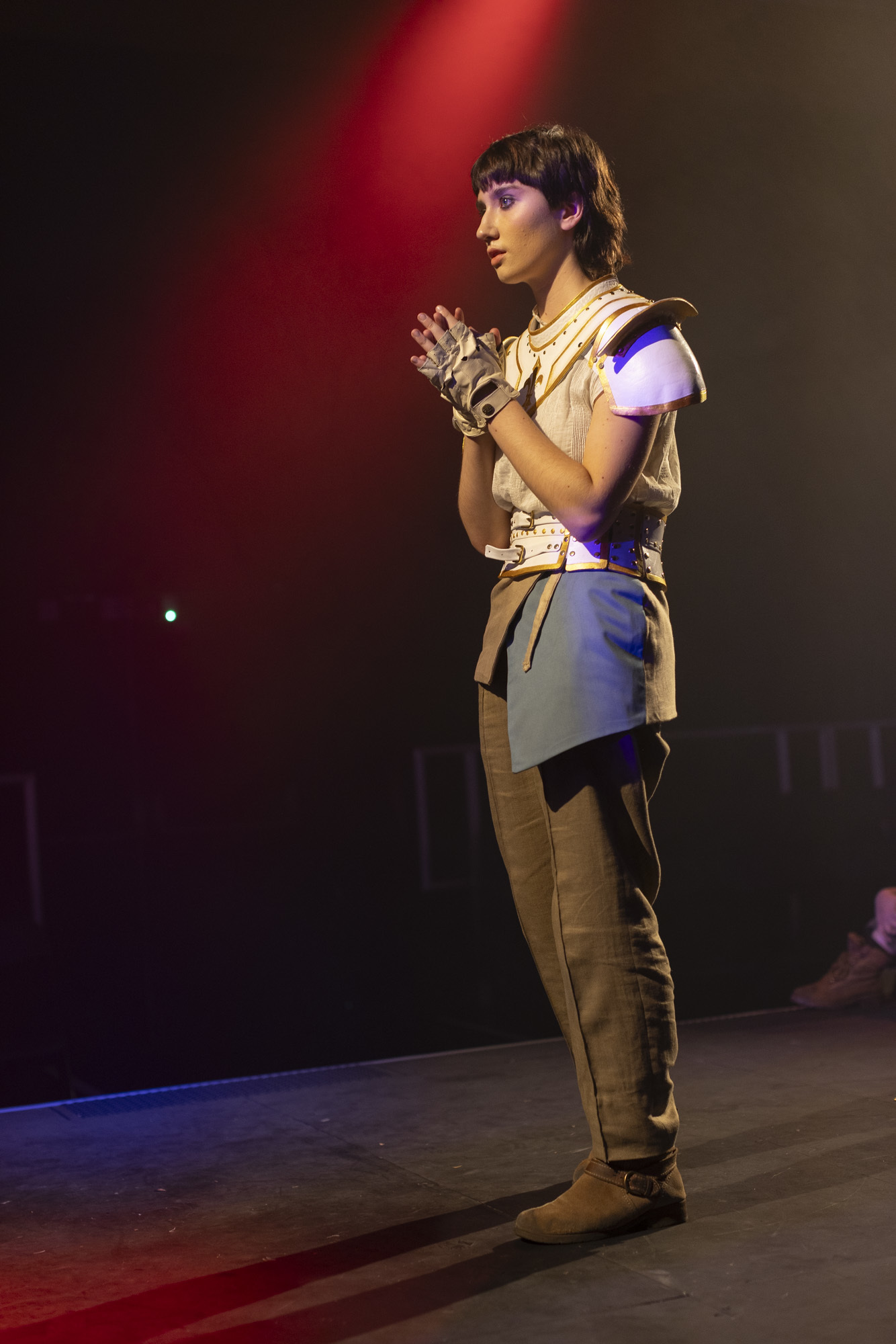 A BA (Hons) Acting student in medieval-style costume and some armour as Joan of Arc. They stand on stage looking straight ahead with a red spotlight shining in the background.