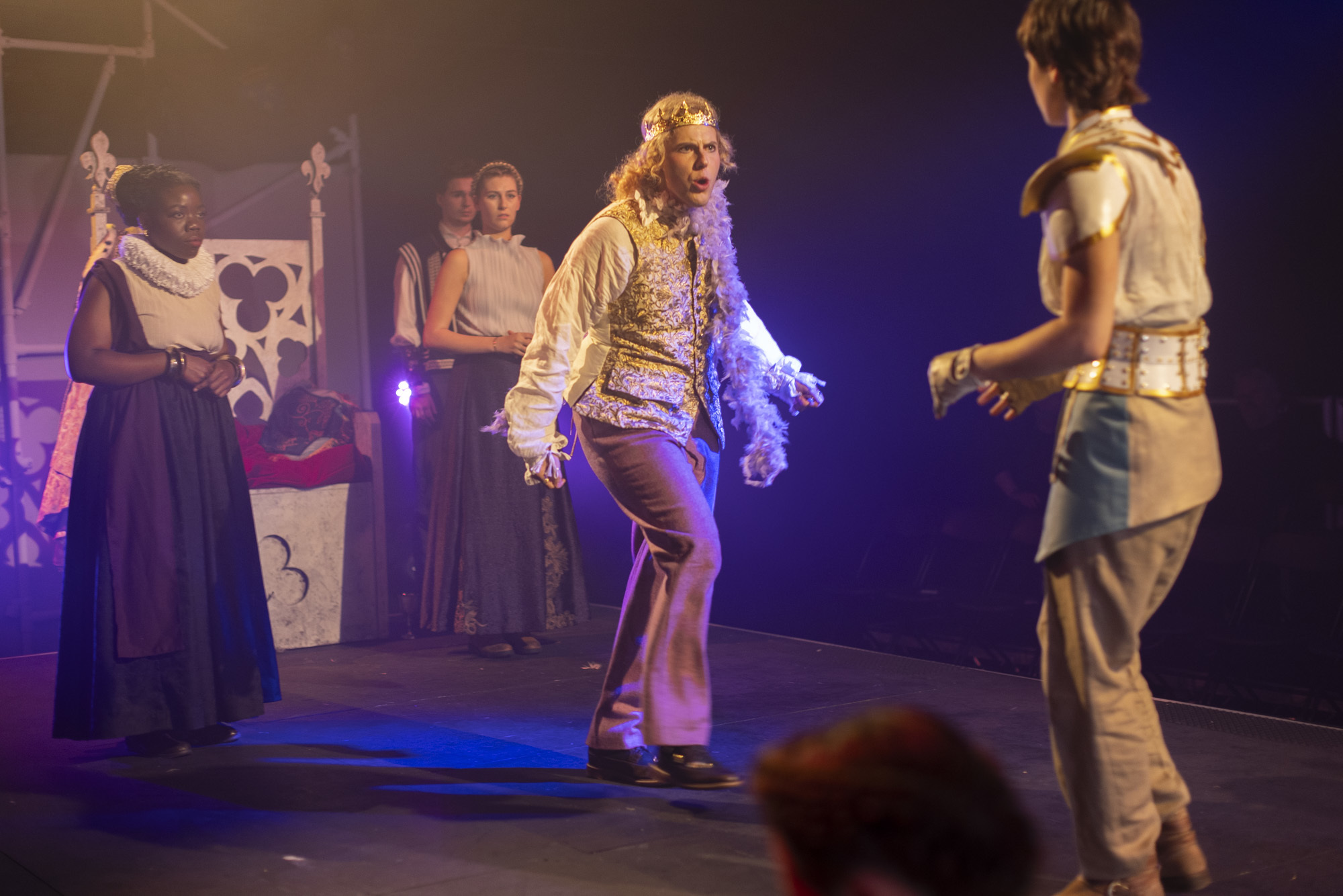 A group of BA (Hons) Acting students performing "I, Joan". One student, dressed as a king, is angrily approaching another student, dressed in some armour as Joan of Arc with their back to the camera. Three other students stand by a throne in the background.