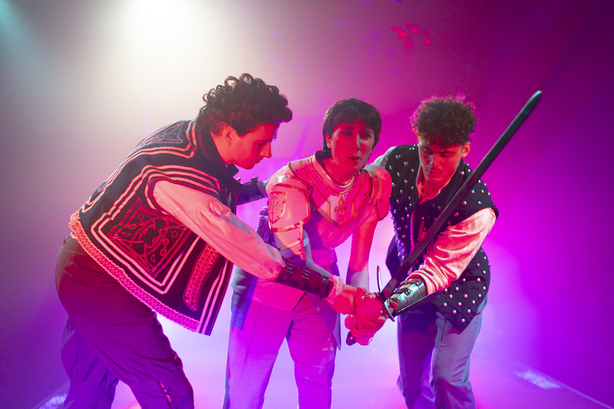 Three BA (Hons) Acting students in costume while performing a production of "I, Joan". All three actors are leaning forward on a purple-lit stage all grasping a sword.