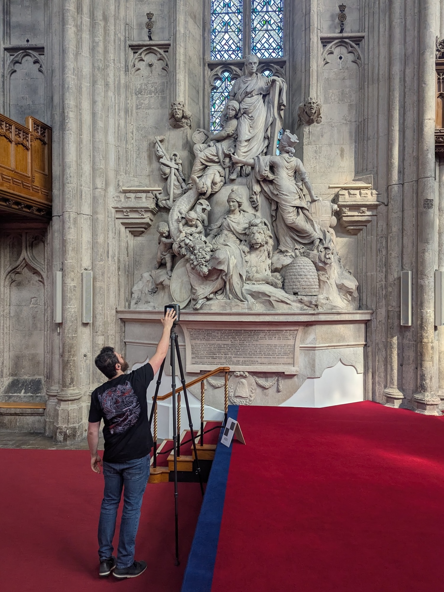 A man stands on a red carpet and reaches towards a large white stone religious sculpture mounted on a cathedral wall, with a stained-glass window above.