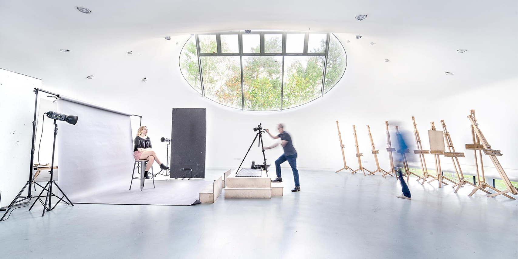 A wide studio space set up for a photo shoot. A model is sitting on a stool  on the left. A photographer is standing behind a tripod and camera mounted on blocks in the centre of the room.