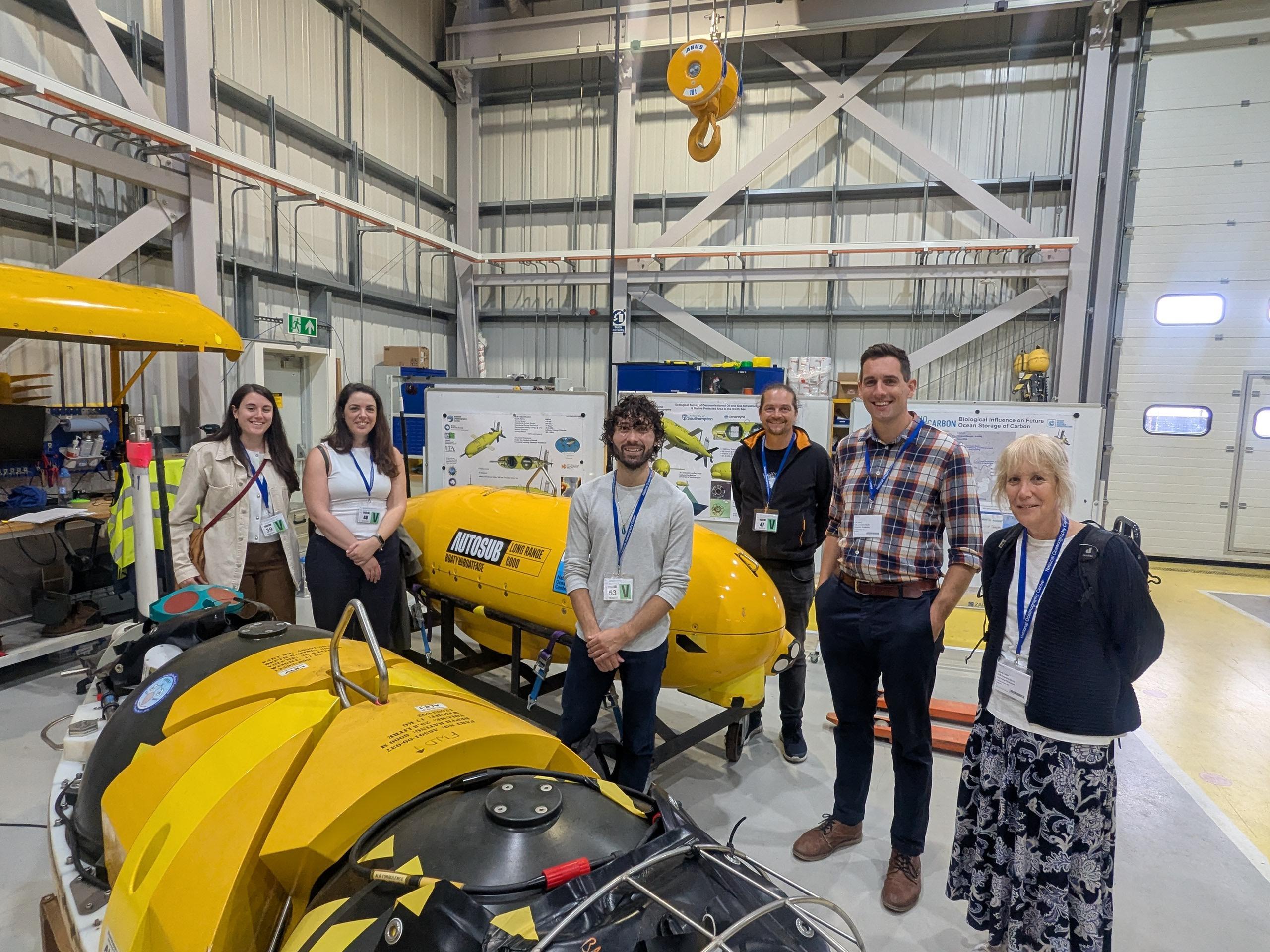 Six people with lanyards stand beside yellow submersible vehicle in industrial warehouse with metal framework and concrete floors.