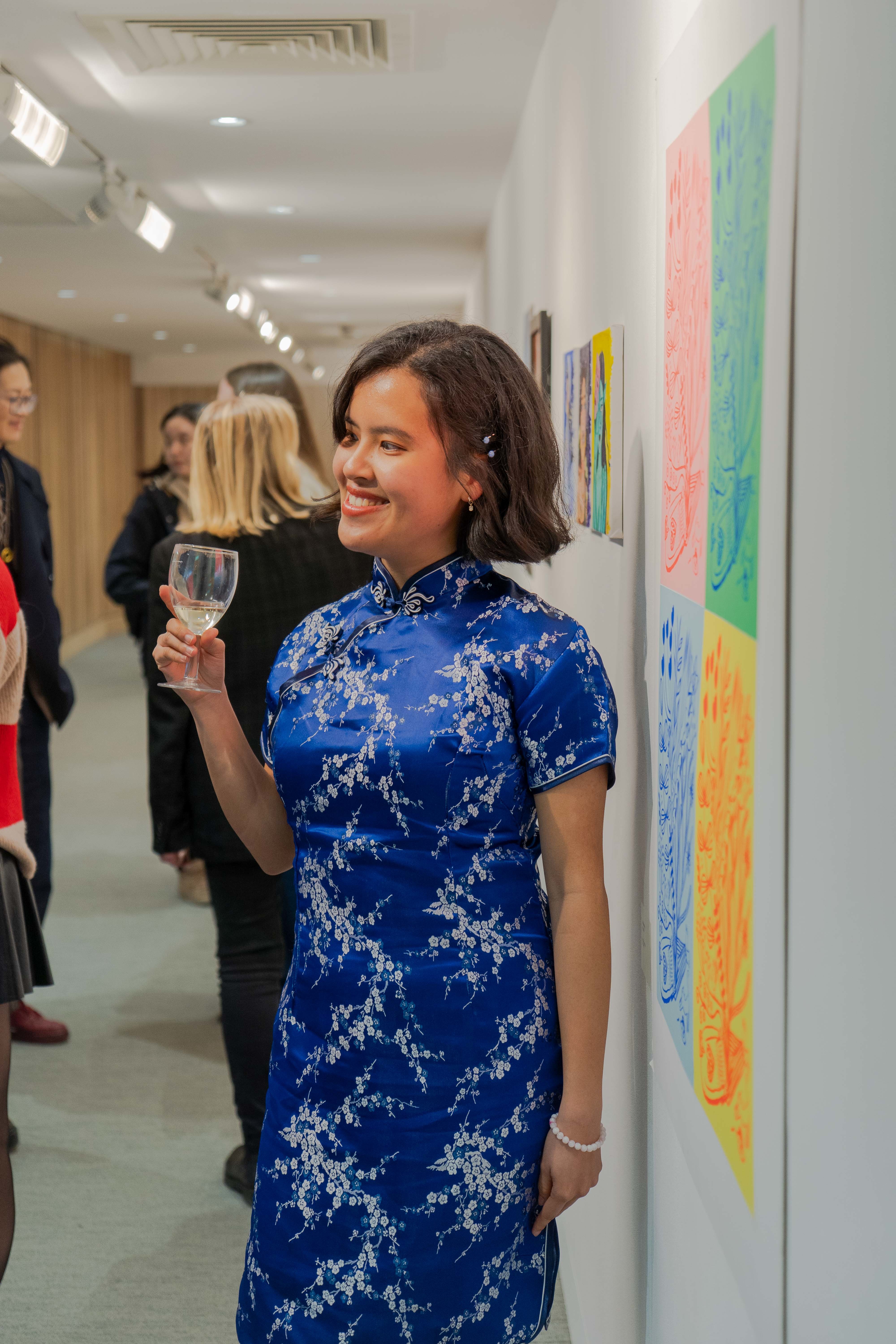 An artist wearing an elegant blue dress smiles and poses next to a colourful abstract artwork at TheGallery, Arts University Bournemouth