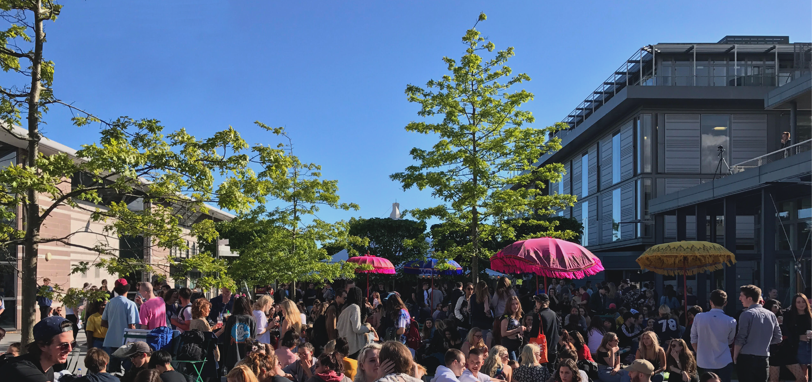 A large crowd of people gather in a courtyard with trees and bright-coloured parasols in the background.