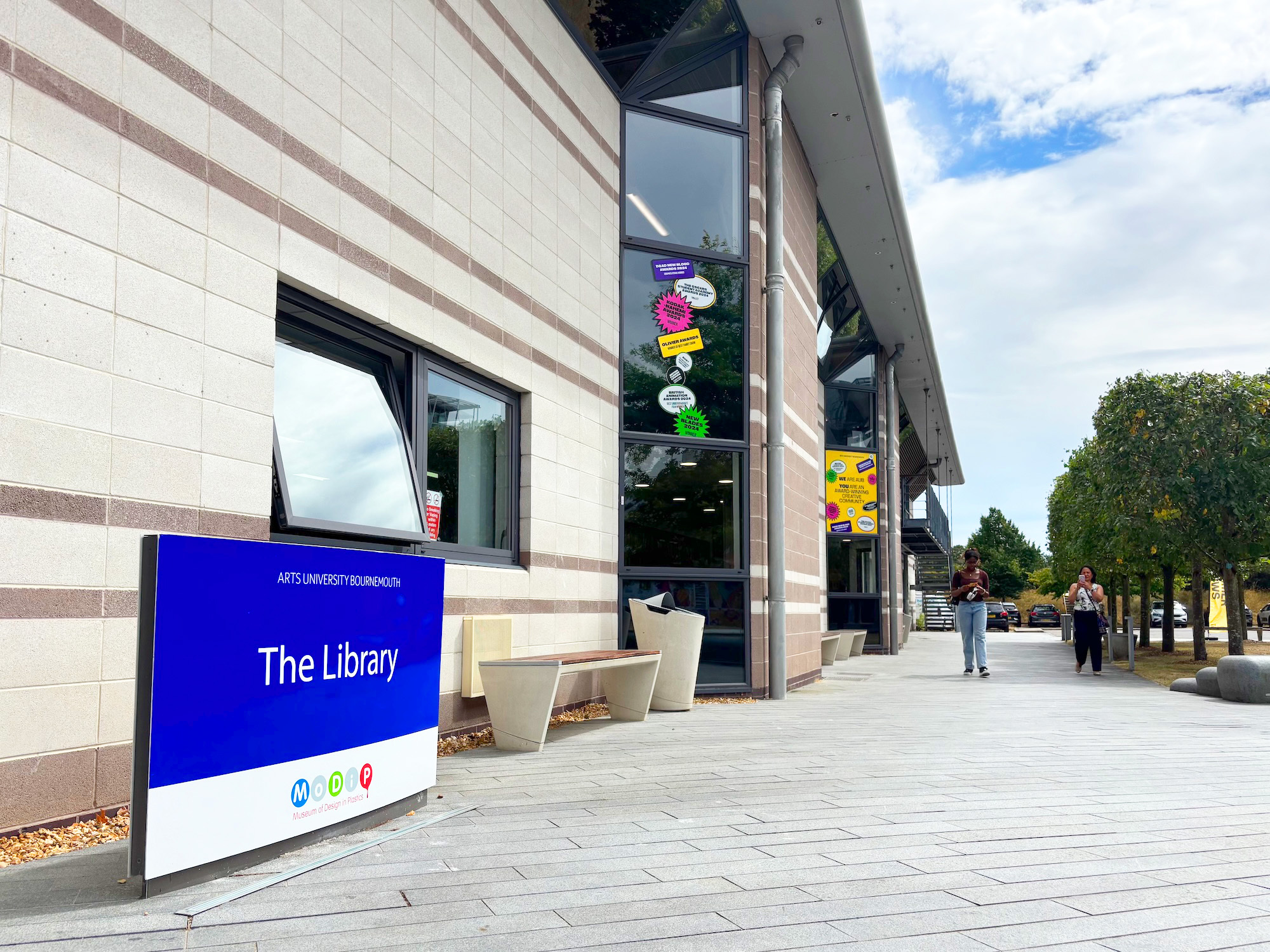 Library at Arts University Bournemouth with glass frontage, blue "The Library" sign, and paved pedestrian area under partly cloudy sky.