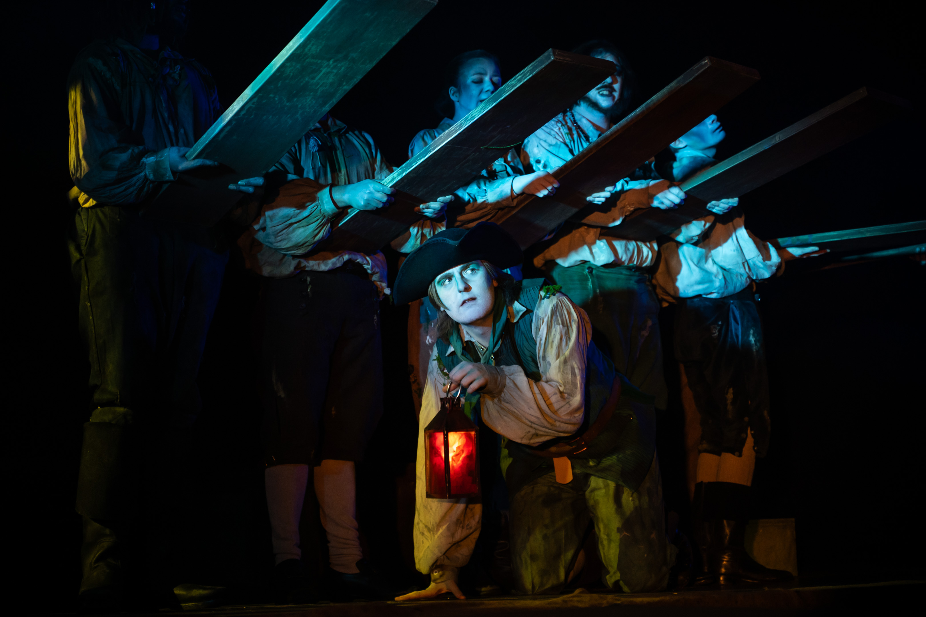 A BA (Hons) Acting student in period costume holding a lantern crawls under a series of planks, held by fellow BA (Hons) Acting students behind them.