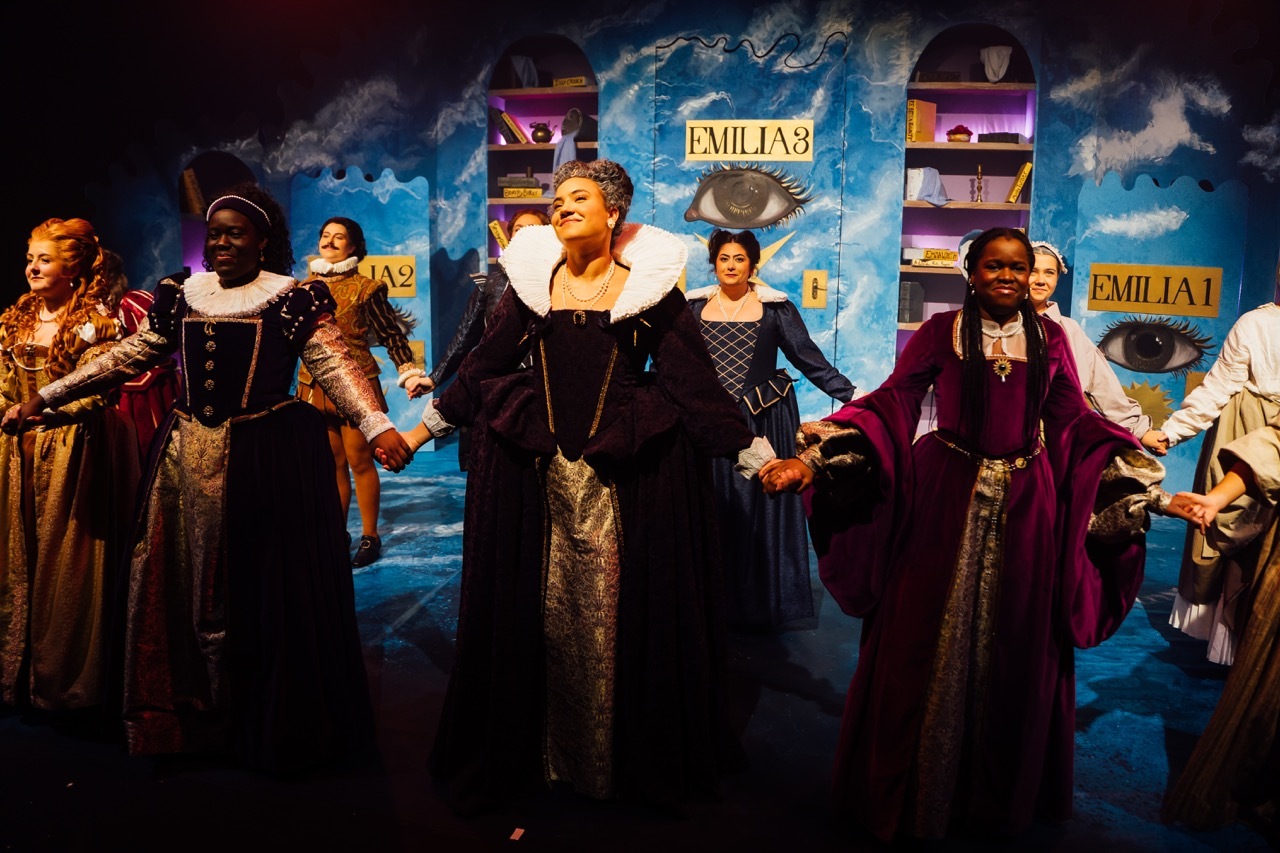 Theatre cast in period costumes taking curtain call on stage, with blue backdrop featuring "EMILIA" text and bookshelves.
