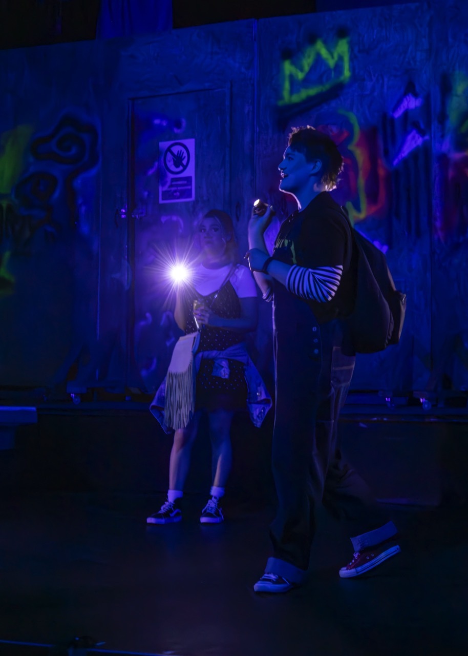 Performer stood next to another in blue and purple light with posters on walls, neon painting in the background.
