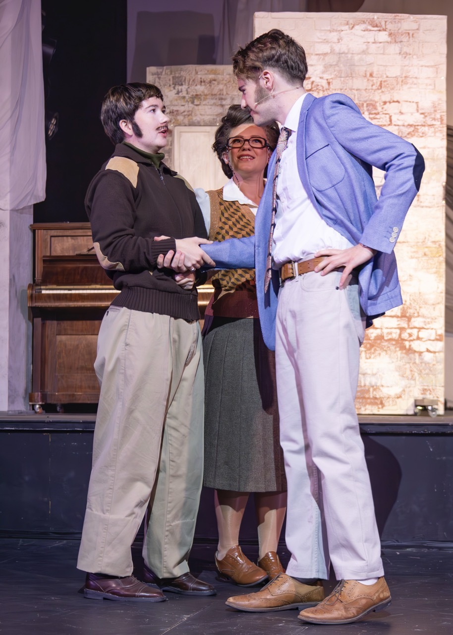 Two men shaking hands, with a woman standing between them, in what appears to be a theatre or performance setting with a brick wall background.