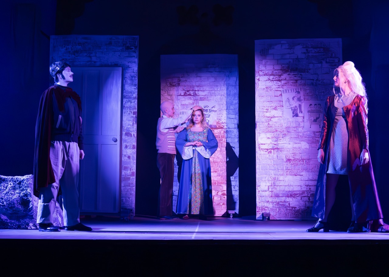 Three actors on stage with brick wall backdrop. Blue and purple lighting illuminates the scene. Left figure in dark costume, centre in blue robe, right in red and black garments.